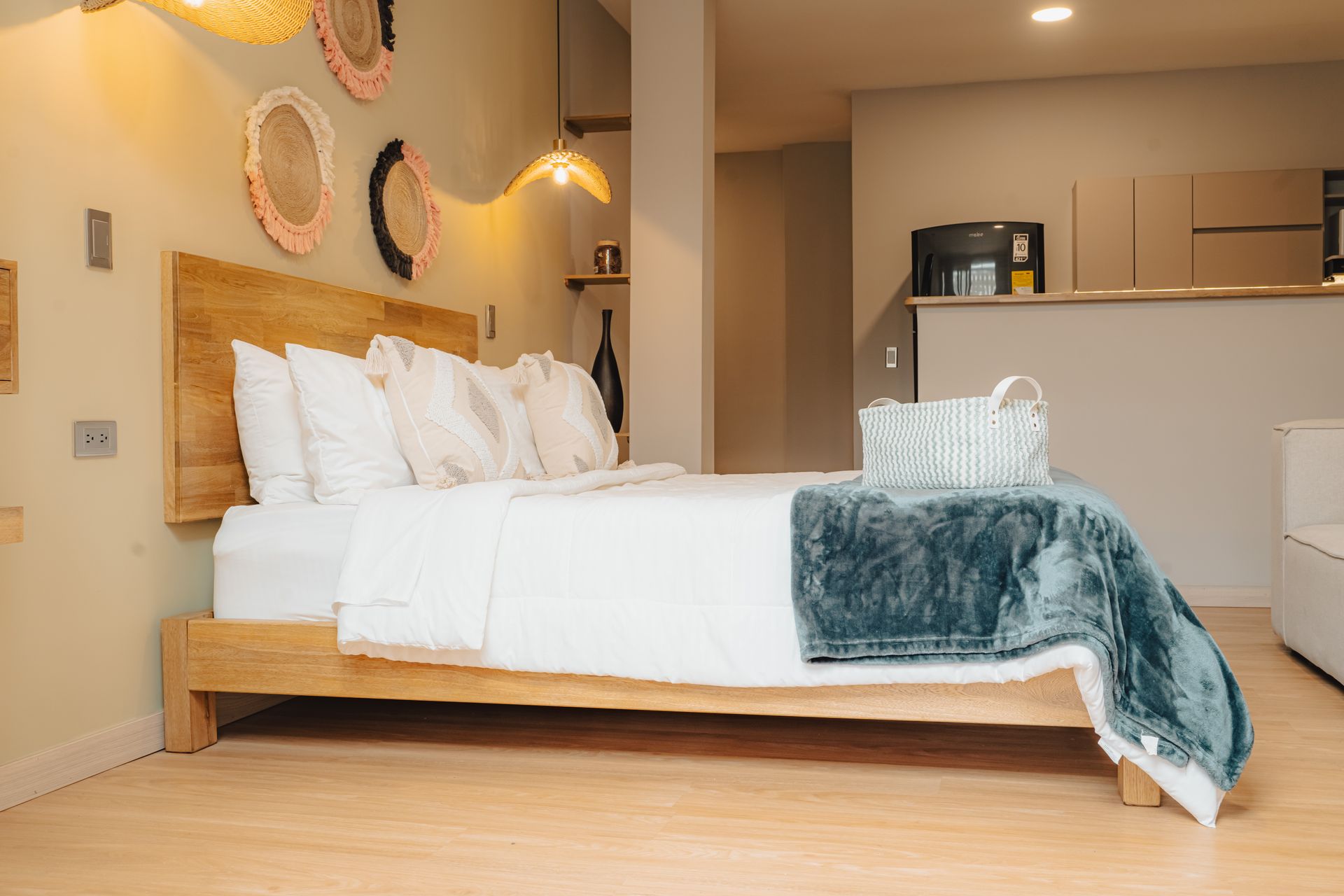 Dormitorio con cama de madera, ropa de cama blanca y manta verde azulado. Tapices decorativos y una pequeña cocina al fondo.