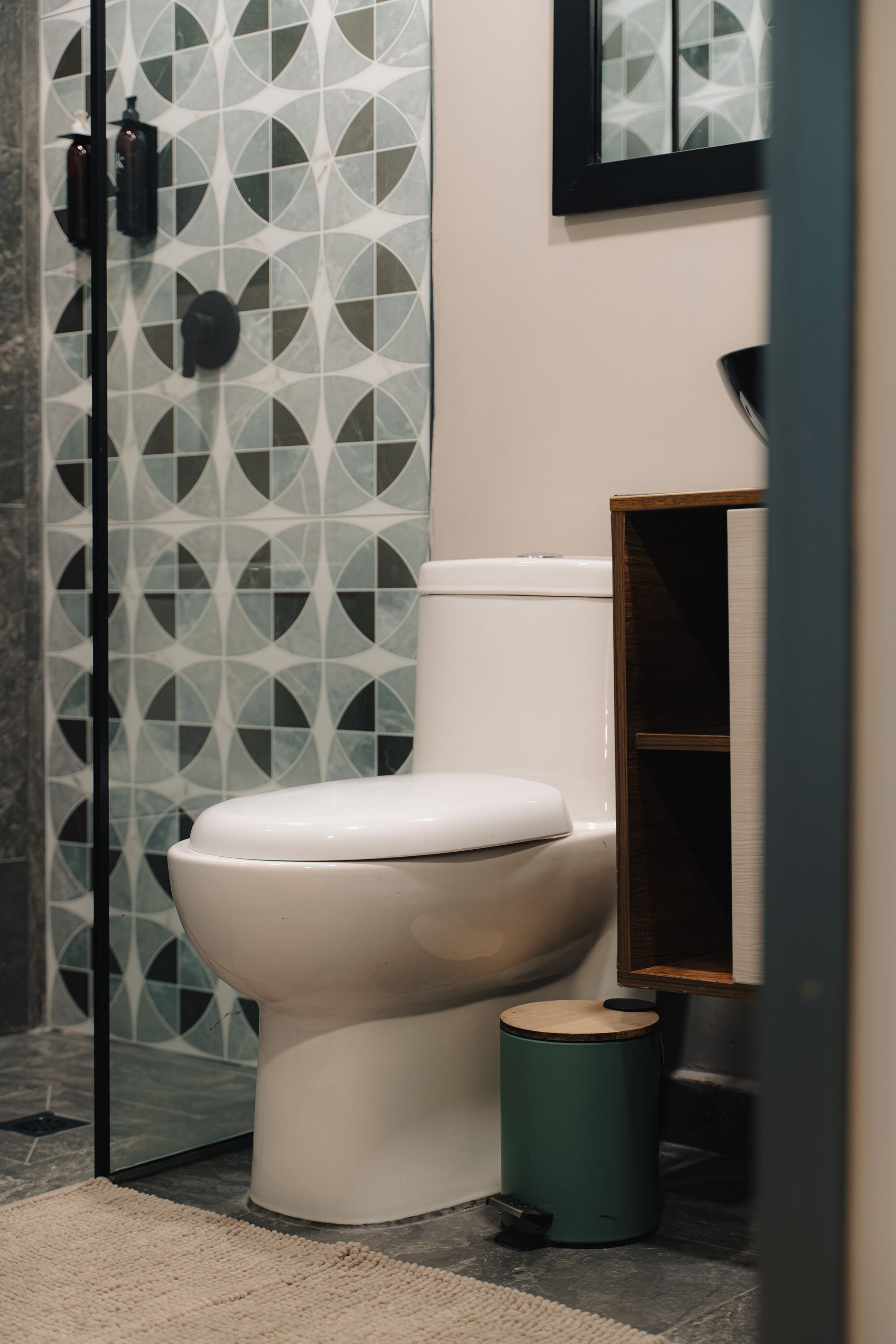 Baño con pared de ducha estampada, inodoro blanco, bote de basura verde y mueble de madera.