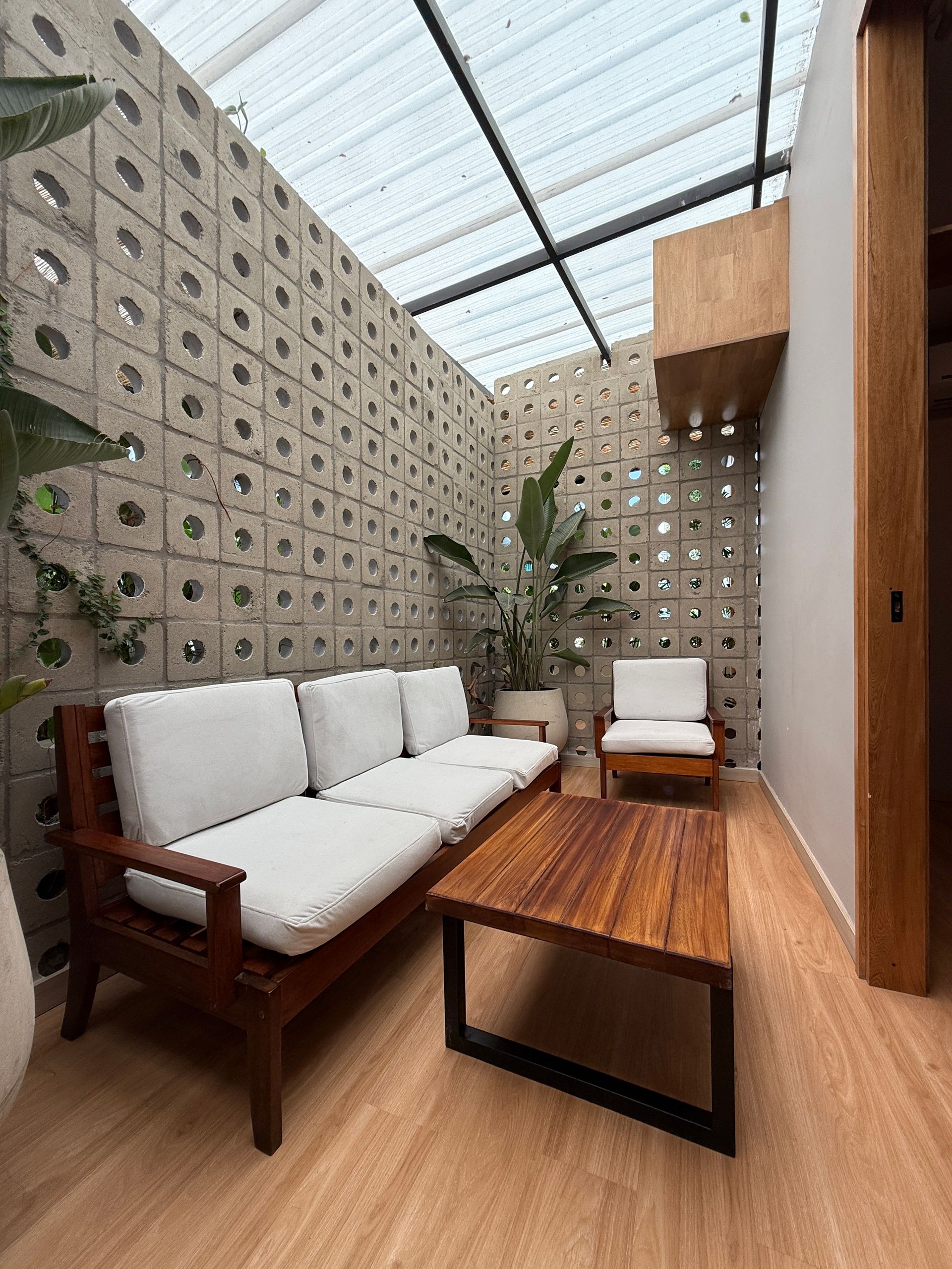 Un patio soleado con sofá, silla y mesa de madera. Paredes de bloques de hormigón y techo transparente.