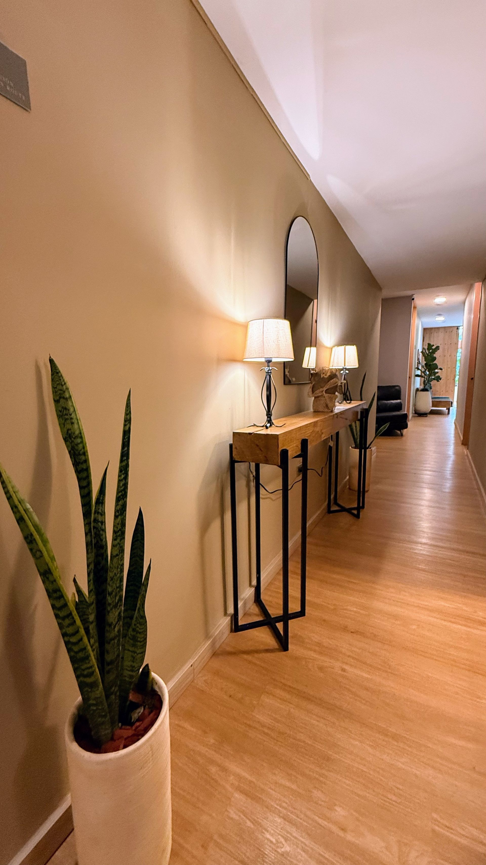 Pasillo largo con suelos de madera clara y paredes color canela. A la izquierda, una consola de madera y lámparas.