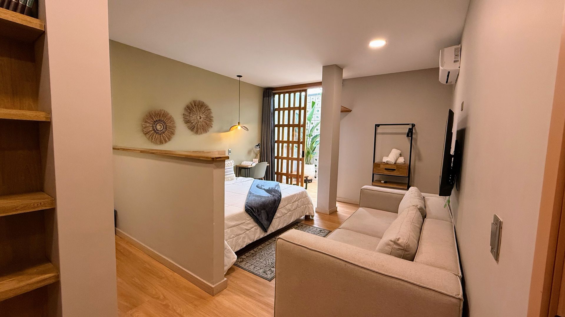 Dormitorio con cama, sofá, estantería y acceso a una zona exterior. Paredes claras y suelo de madera.