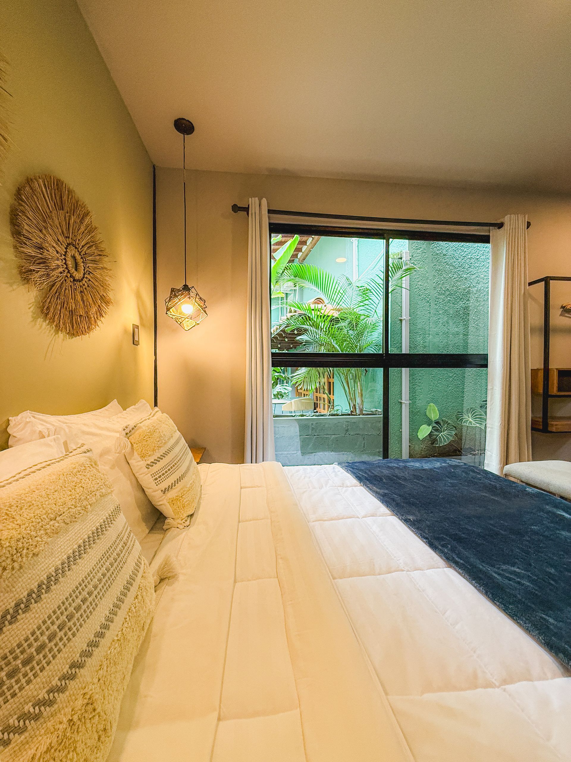 Dormitorio con cama, manta azul y vistas a la vegetación a través de una ventana. Paredes amarillas, lámparas colgantes.