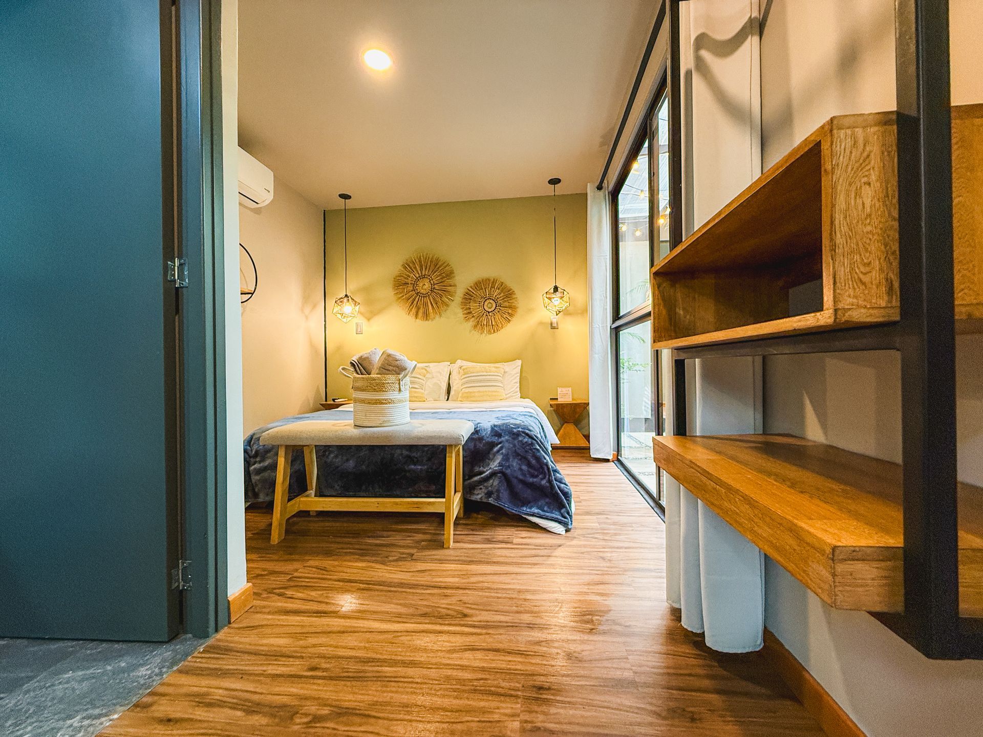 Interior de dormitorio con cama, estanterías y grandes ventanales. Suelos de madera cálidos y paredes verde claro.