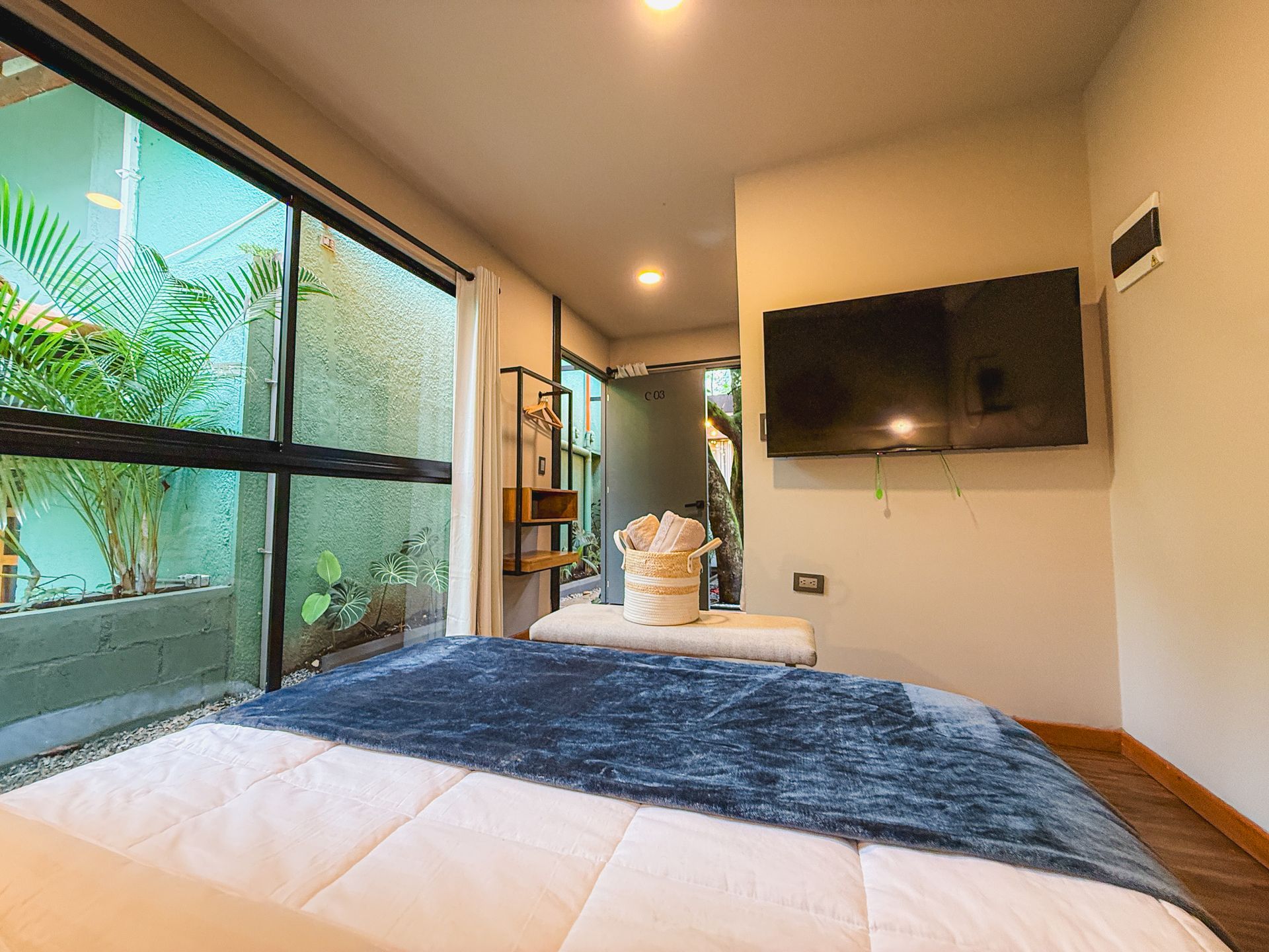 Dormitorio con ventana grande, cama y televisor. Manta azul, paredes claras y vista al exterior.