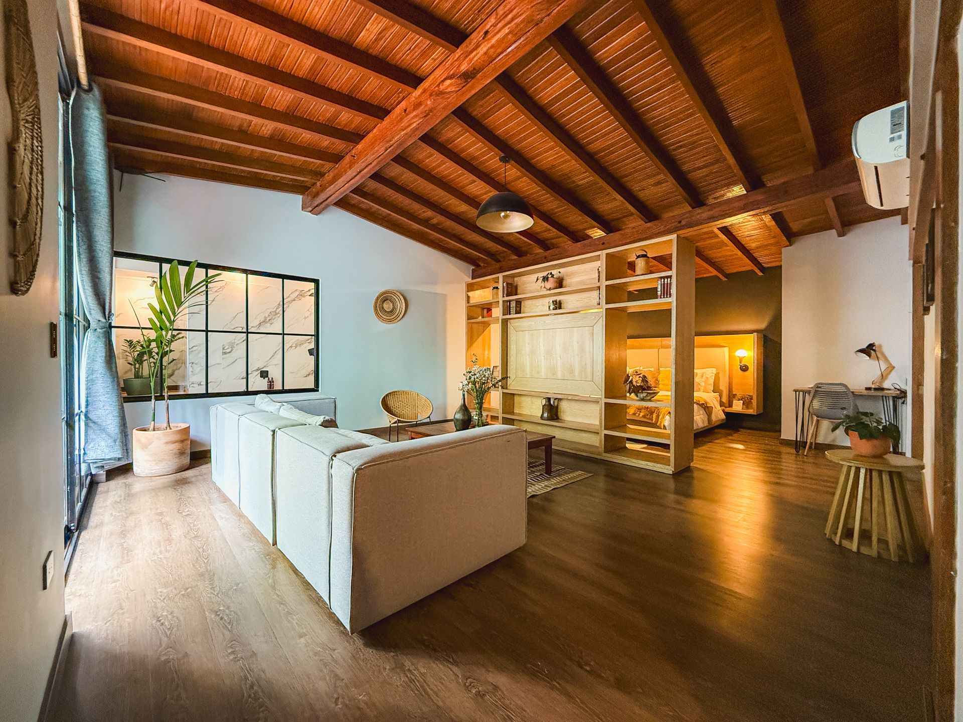 Sala de estar con techo y piso de madera, sofá blanco, estanterías empotradas, gran ventanal.