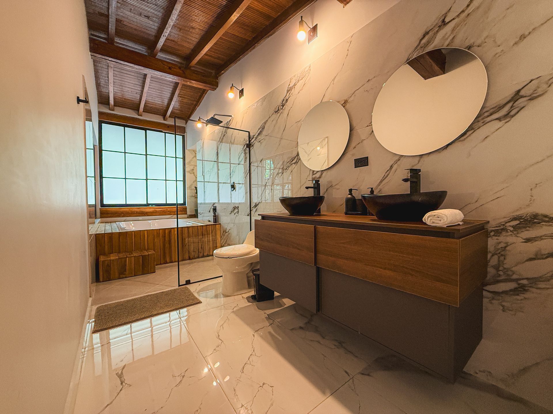 Baño con paredes de mármol, tocador de madera, lavabos negros, espejos redondos y ducha de cristal.