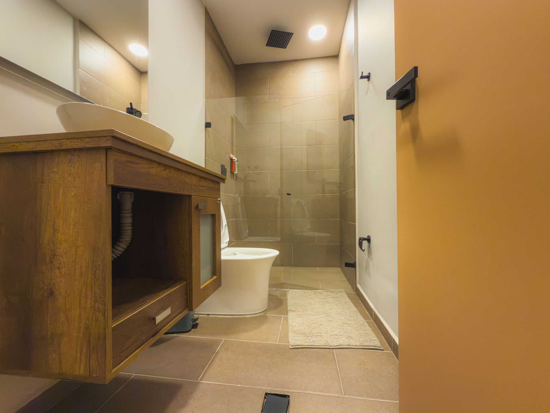 Baño con tocador de madera, lavabo e inodoro blancos, ducha de cristal y puerta abierta.