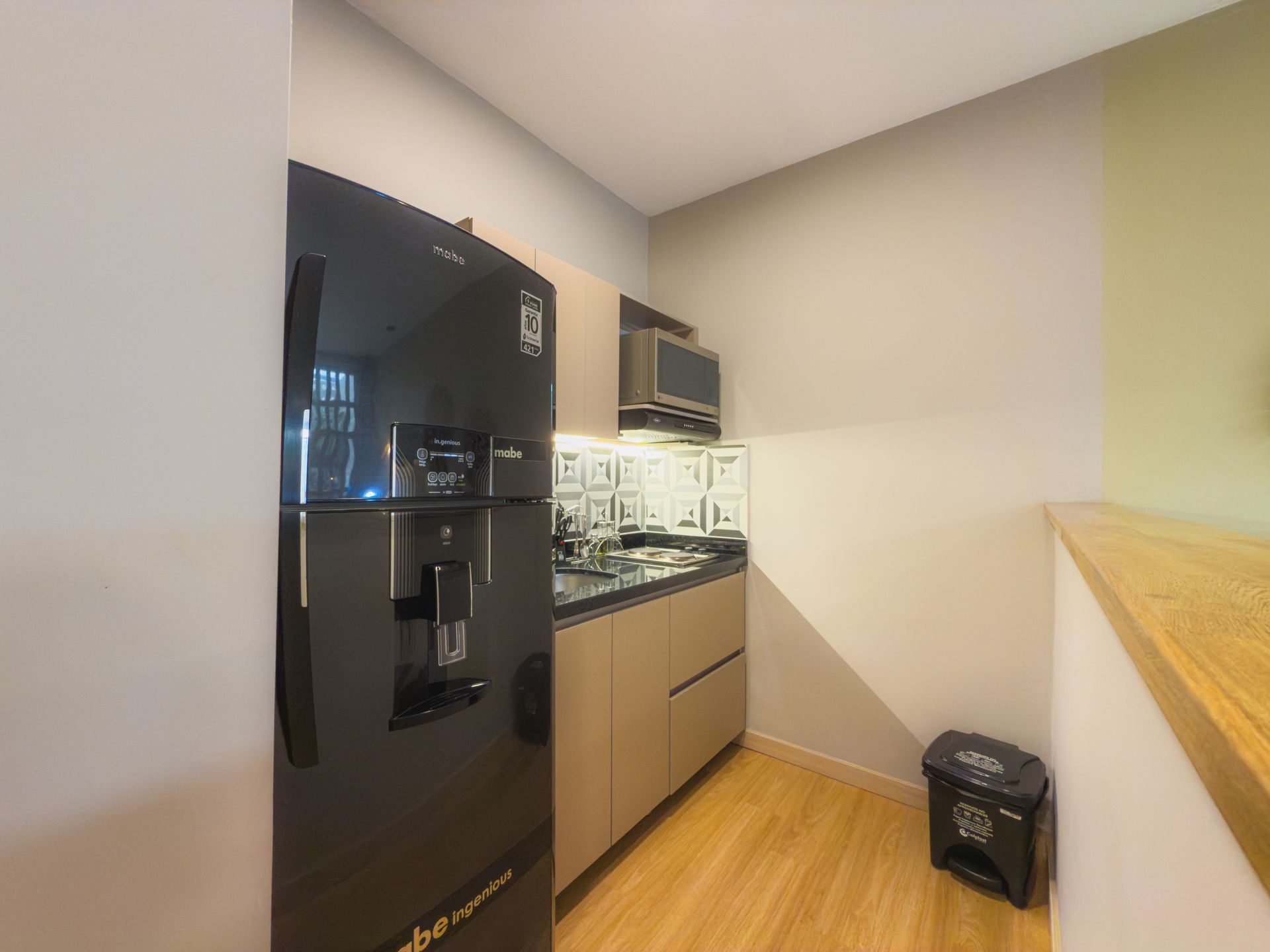 Cocina pequeña con refrigerador negro, microondas y gabinetes beige; hay un bote de basura cerca.