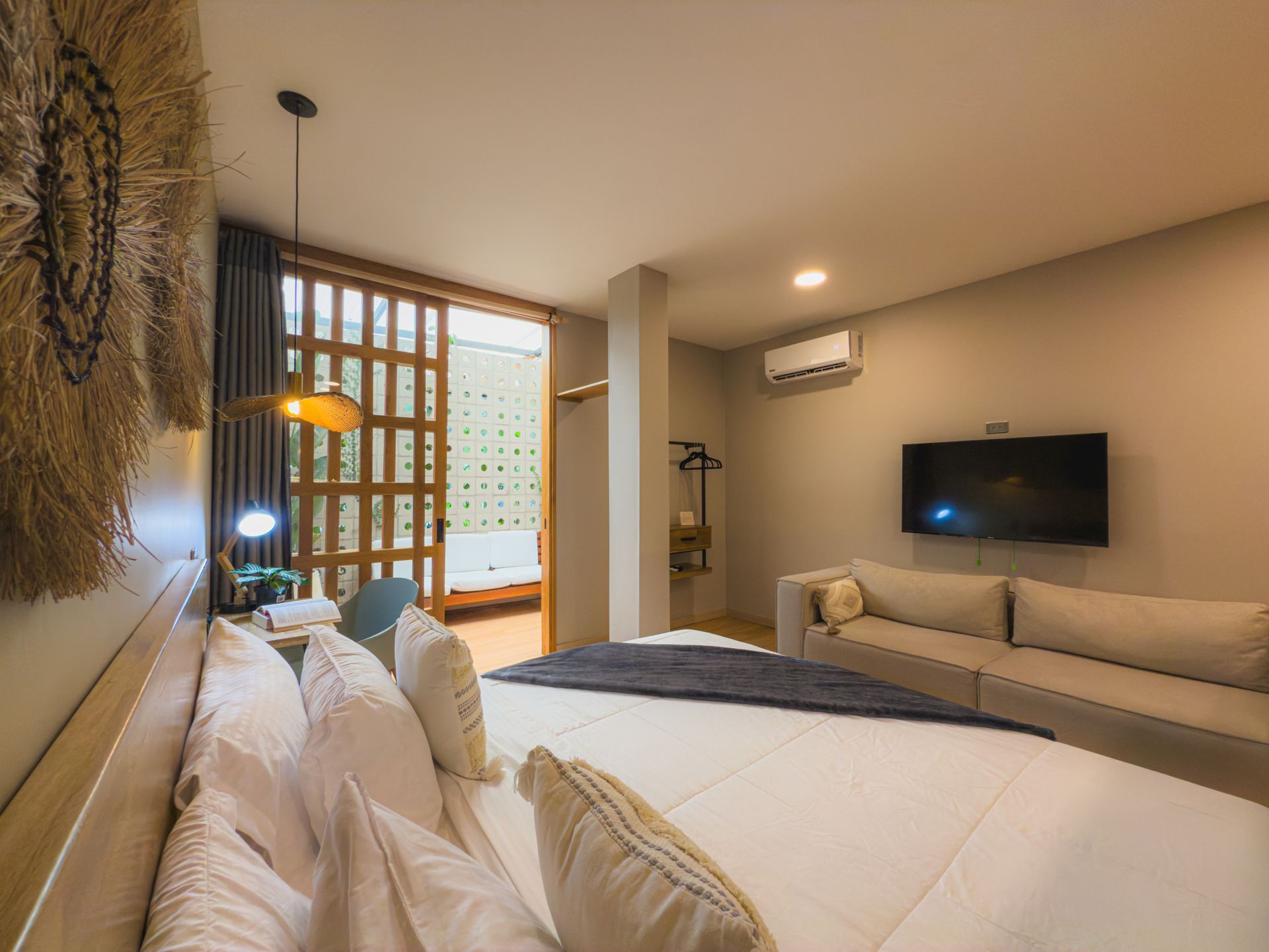 Dormitorio con cama grande, sofá, TV y puerta a un balcón. Tonos beige y blanco.