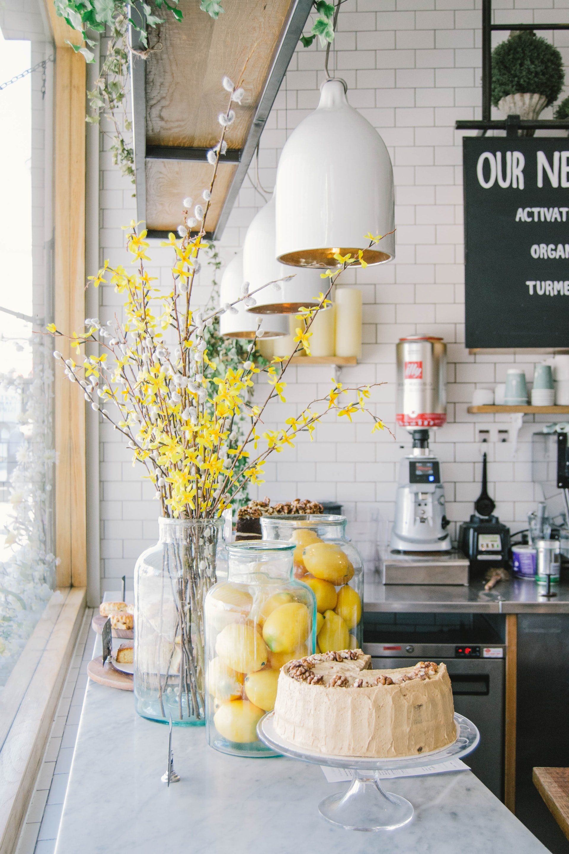 Un luminoso mostrador de cafetería con flores amarillas, limones y un pastel. Lámparas colgantes blancas cuelgan del techo.