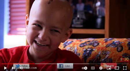 Smiling bald child in a red shirt, sitting on a couch with a patterned blanket behind, in a video player view