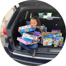 Child sitting in an SUV trunk, smiling and holding stacked toy boxes and colorful packages