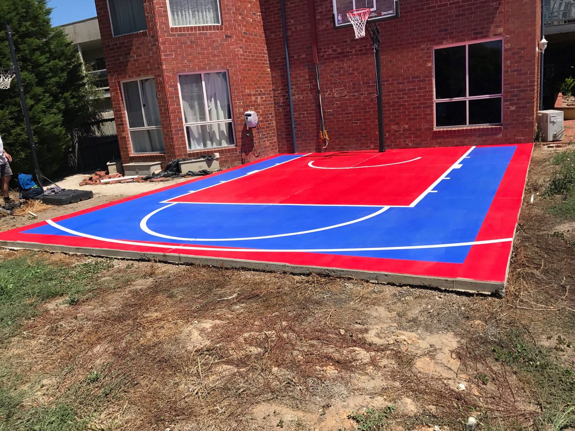 A red and blue basketball court is being built in front of a brick house.