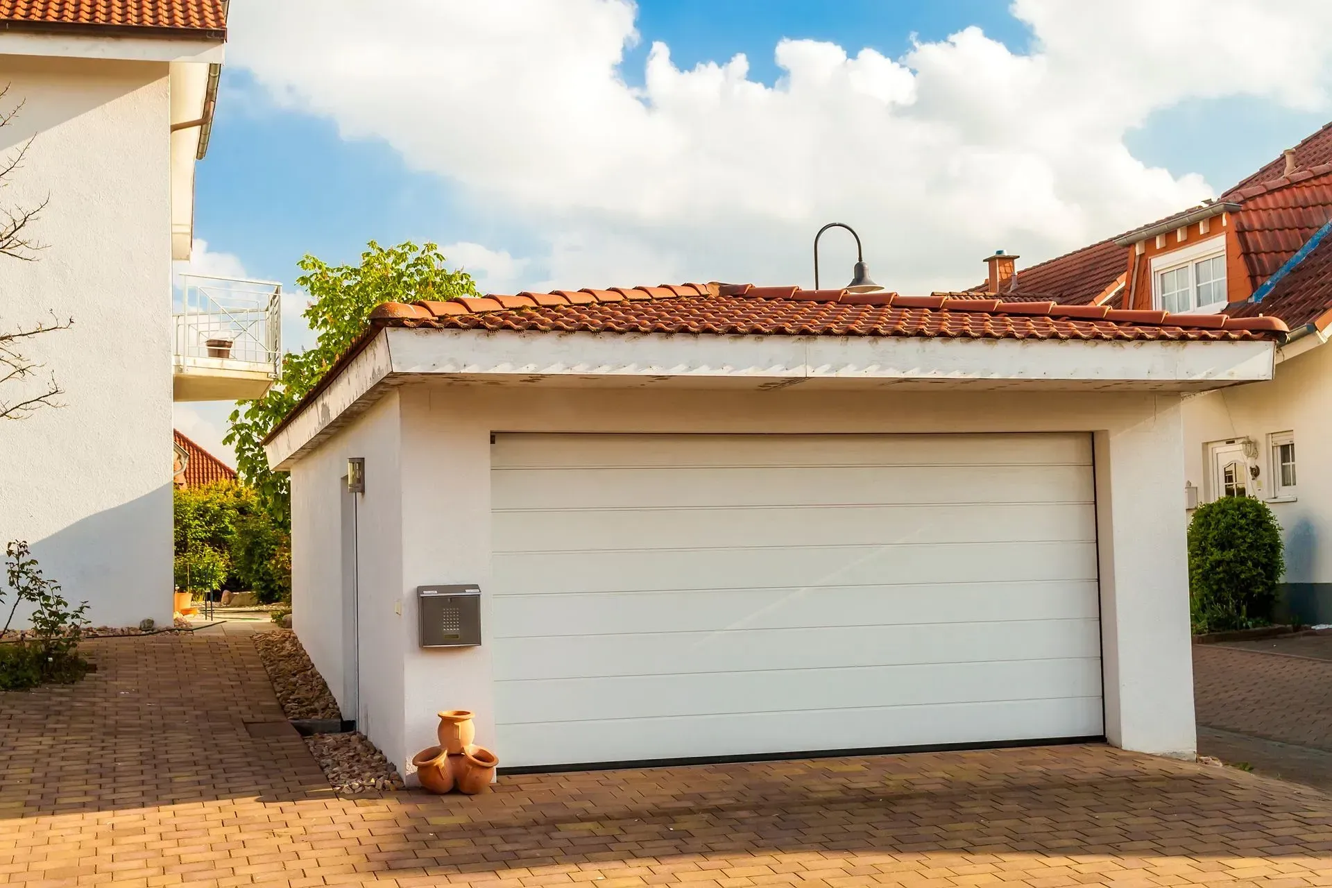White garage with closed door, brown tile roof, next to a white house, on a sunny day.