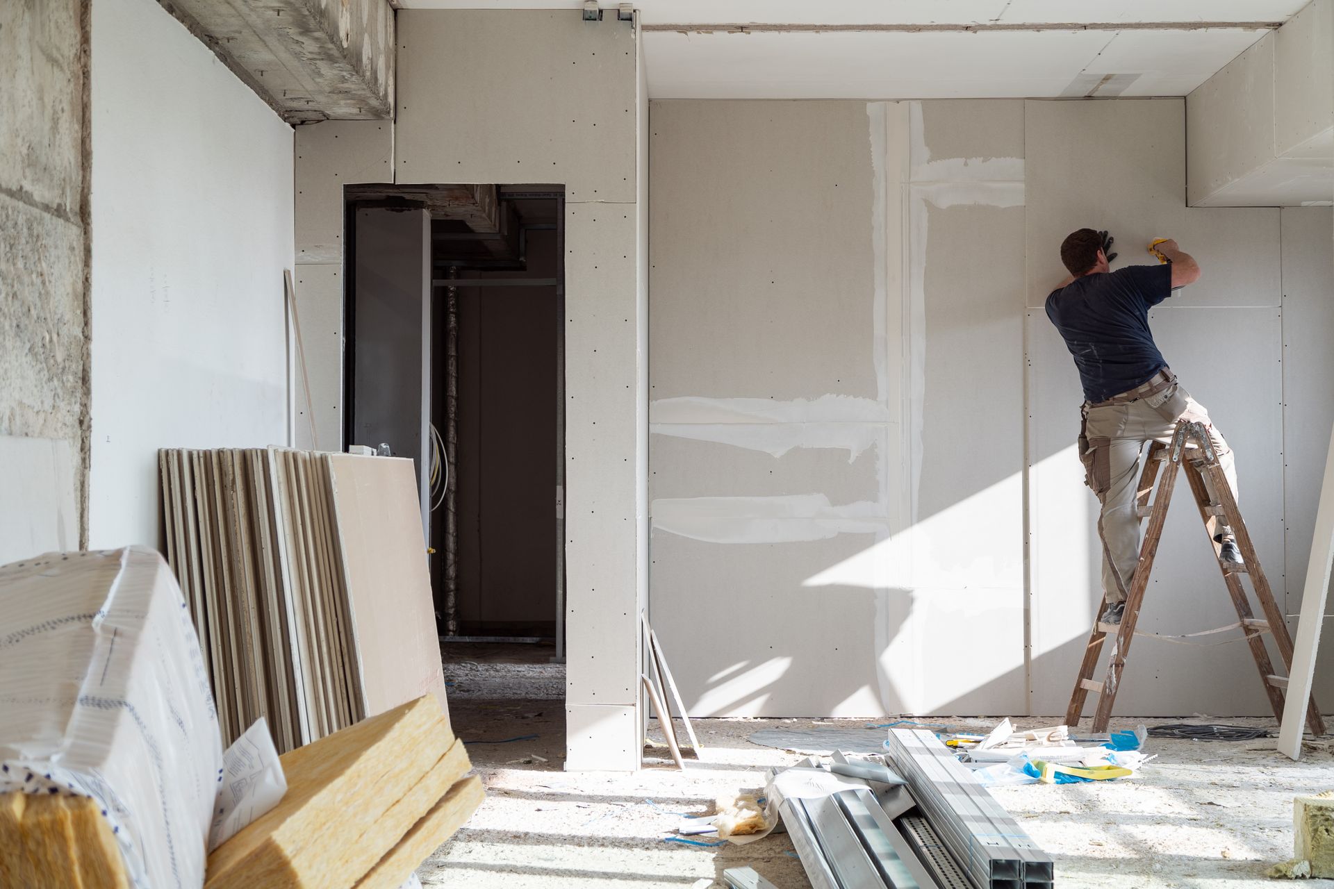 Construction worker on a ladder installing drywall in a building interior. Materials and tools scattered around.
