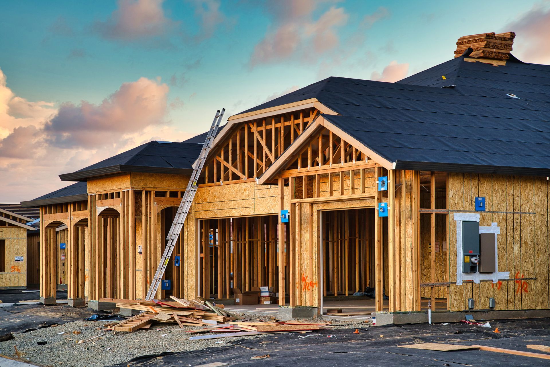 House under construction, wooden frame, dark roof, ladder propped against the structure, blue sky.