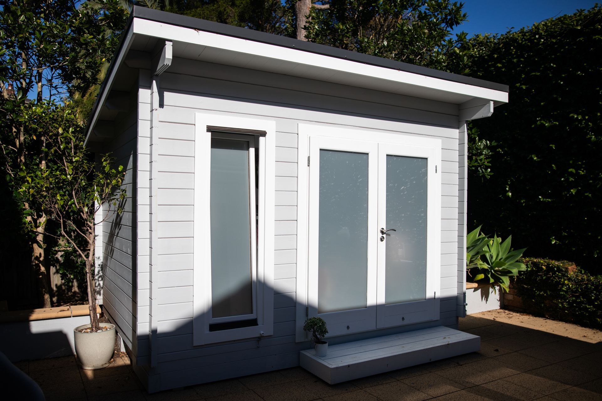 Small gray shed with frosted glass doors and window. White trim, steps, and dark roof.