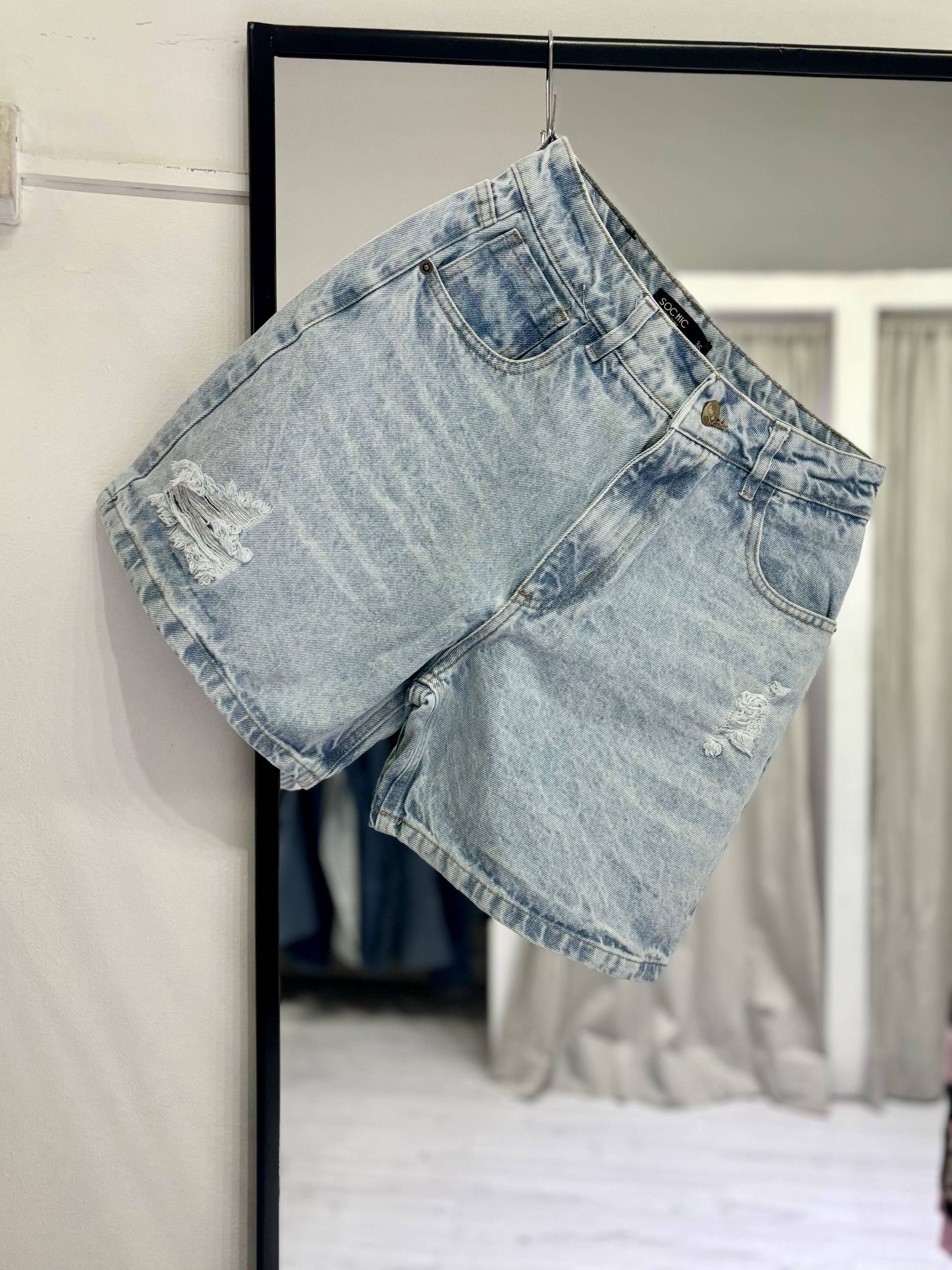 Unos pantalones cortos vaqueros de lavado claro con detalles desgastados cuelgan delante de un espejo.