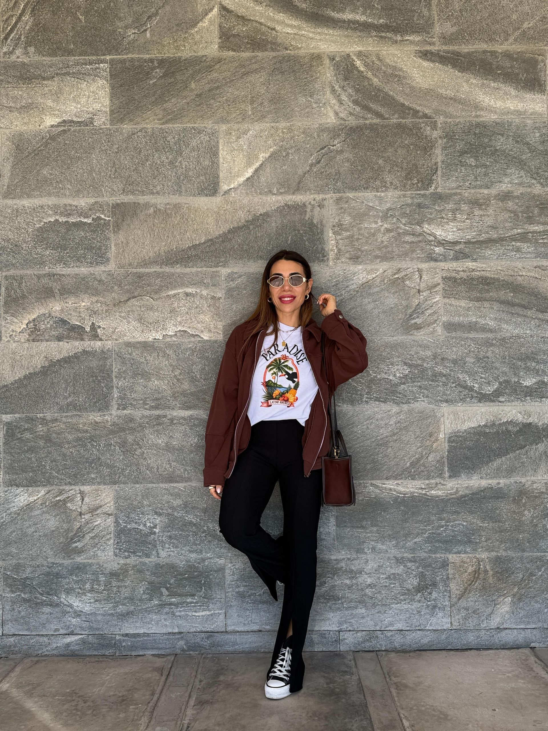 Una mujer con chaqueta marrón y camiseta estampada se apoya contra una pared de piedra gris.