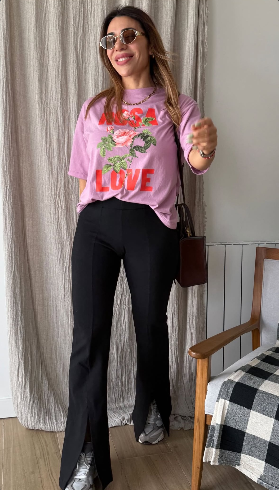 Mujer con chaqueta marrón y pantalones negros, apoyada contra una pared de piedra gris, con gafas de sol.