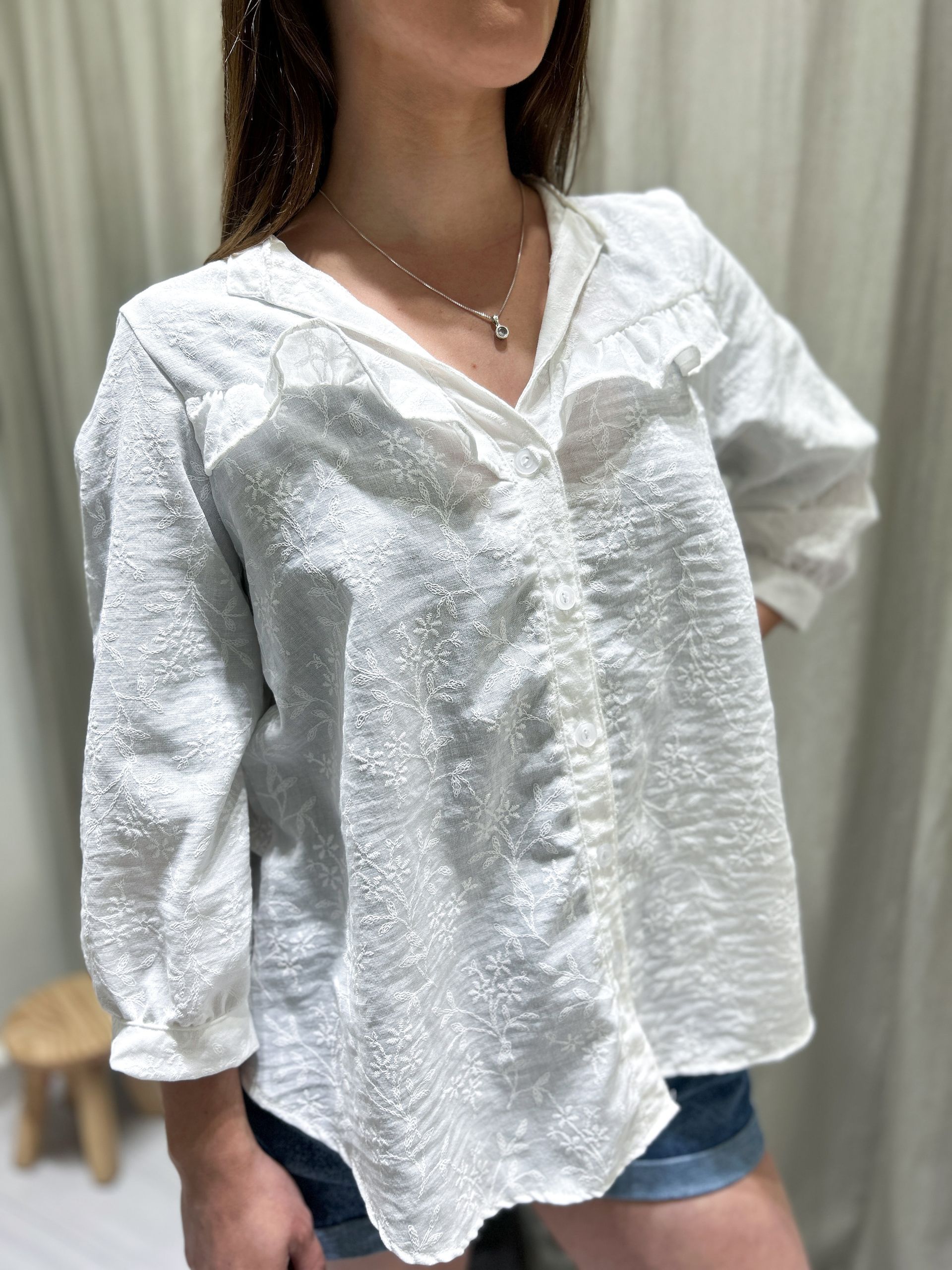 Mujer con camisa blanca abotonada de textura con detalles de volantes; se ven pantalones cortos vaqueros.
