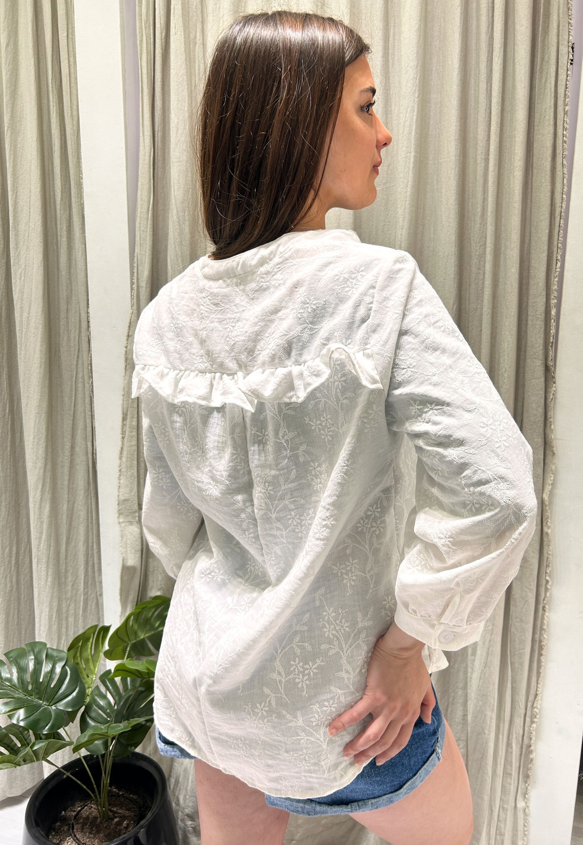 Mujer con camisa blanca abotonada de textura con detalles de volantes; se ven pantalones cortos vaqueros.