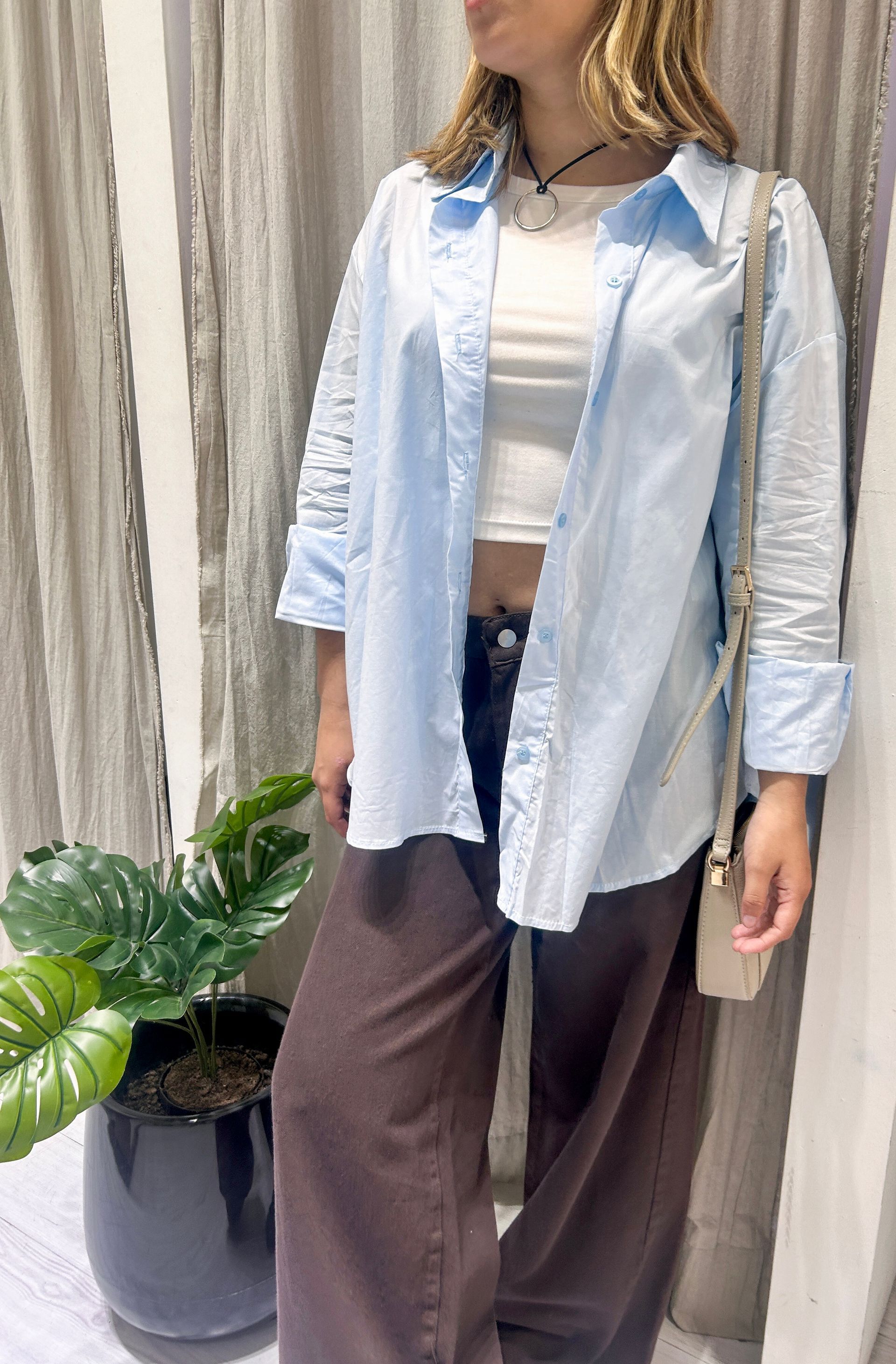 Mujer con camisa azul claro y pantalones marrones, sosteniendo un bolso, cerca de una planta.