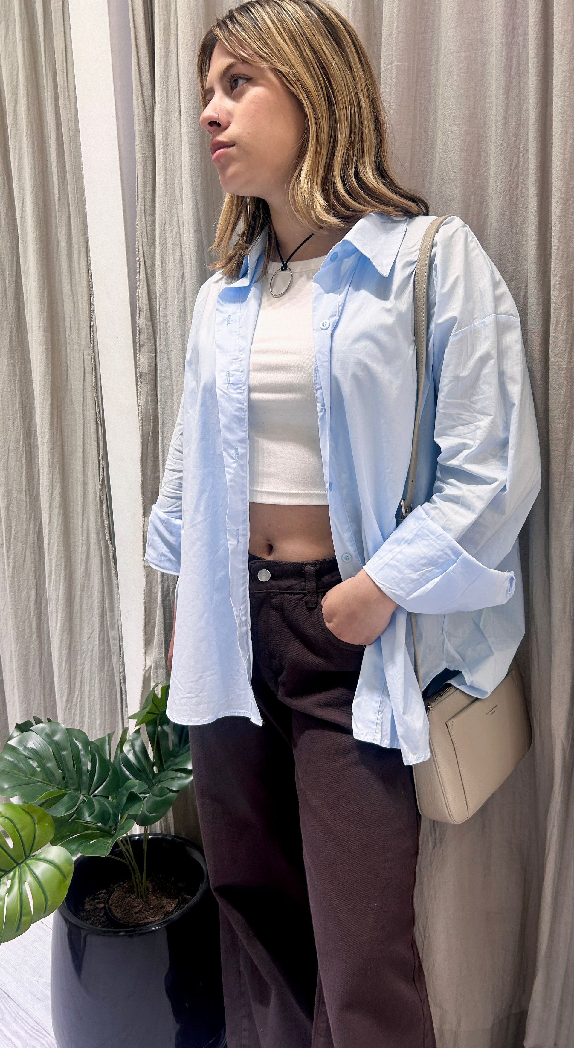 Mujer con camisa azul claro y pantalones marrones, sosteniendo un bolso, cerca de una planta.