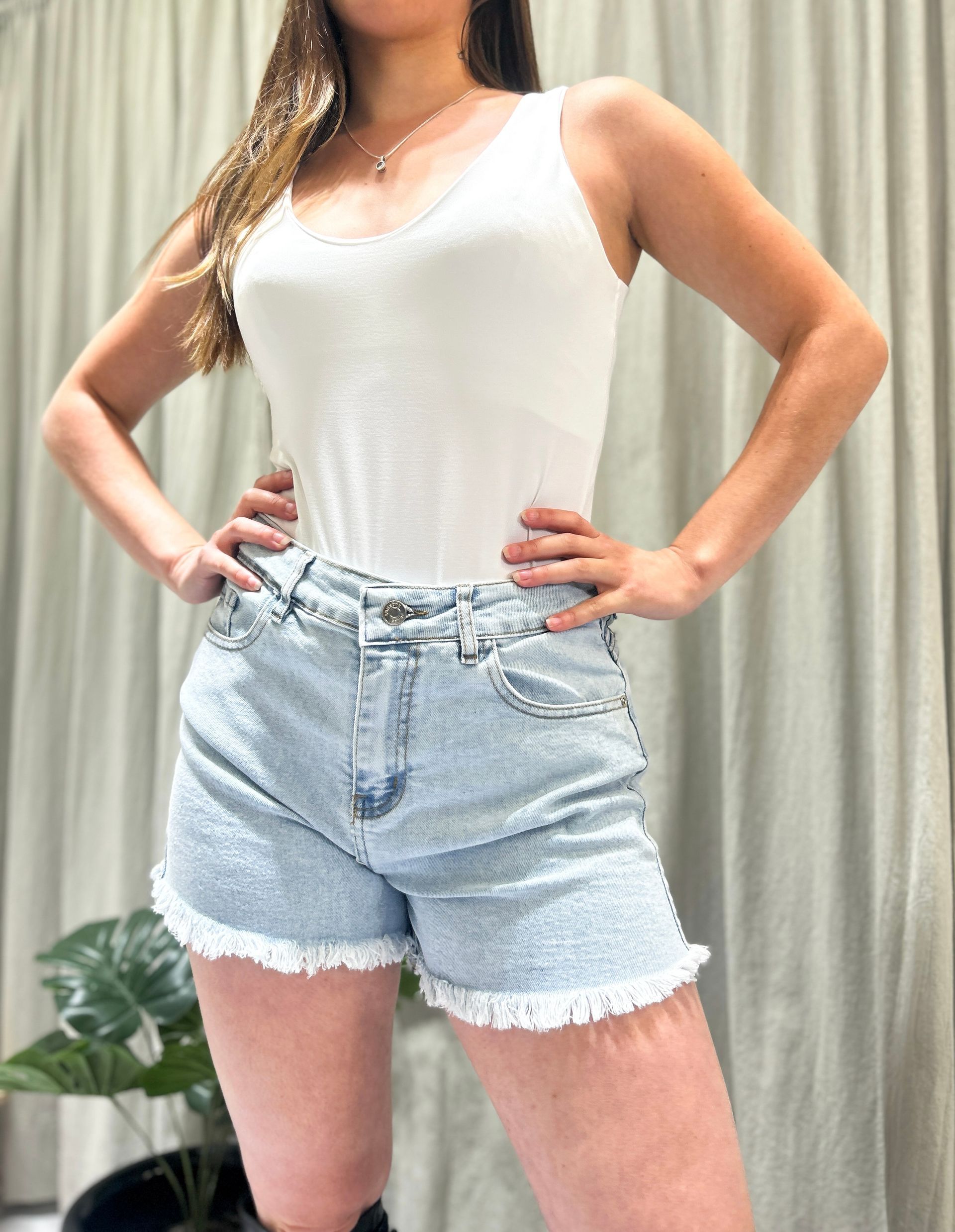 Mujer con camiseta blanca de tirantes y pantalones cortos vaqueros claros, posando con las manos en las caderas.