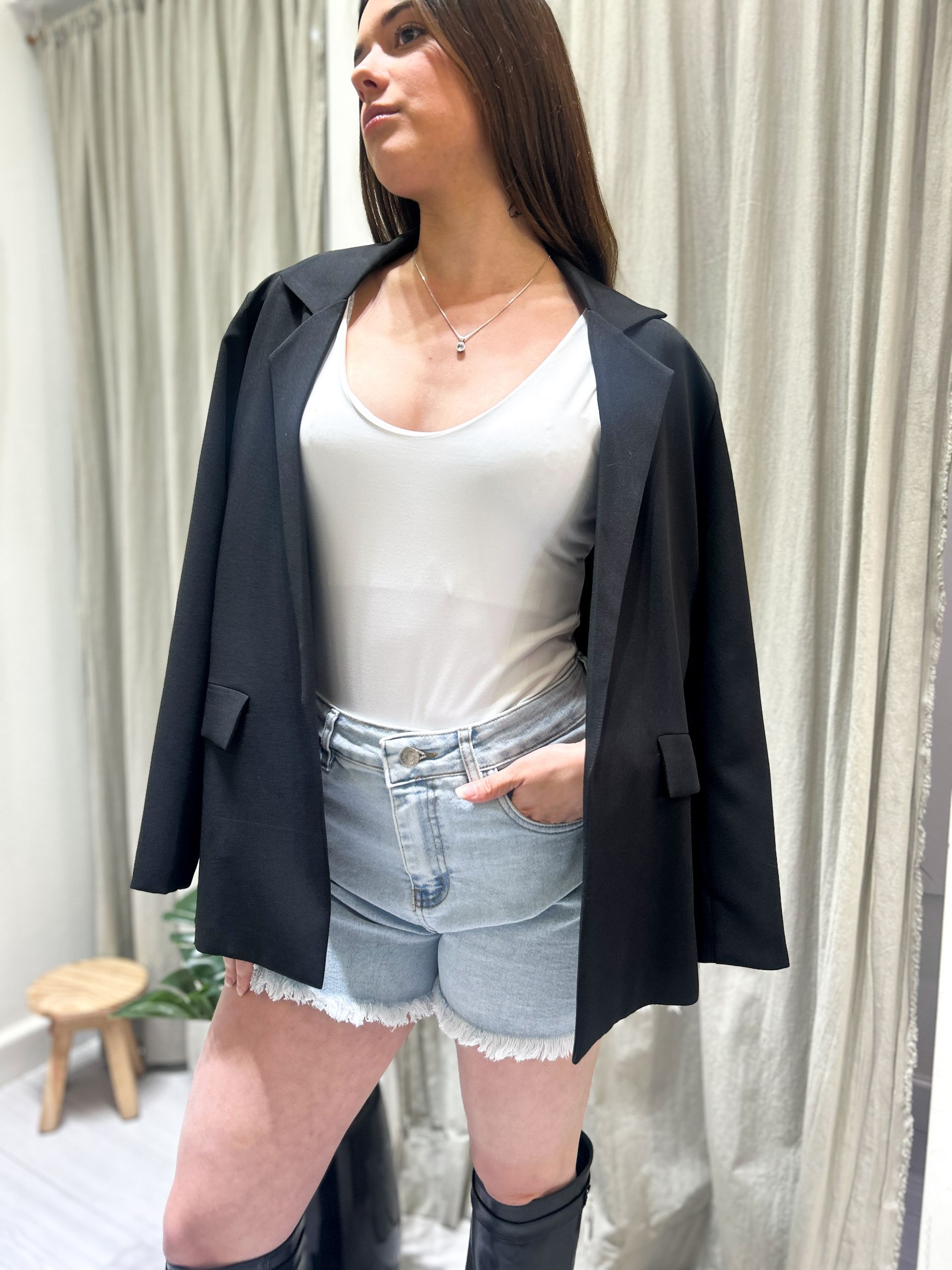 Mujer con blazer negro, blusa blanca, shorts vaqueros y botas en un entorno comercial.