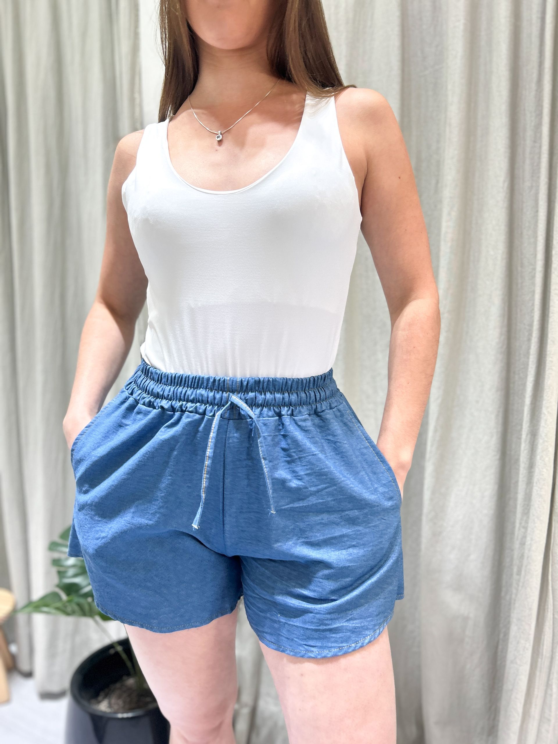 Mujer vestida con una camiseta blanca de tirantes y pantalones cortos azules, con las manos en los bolsillos, de pie.