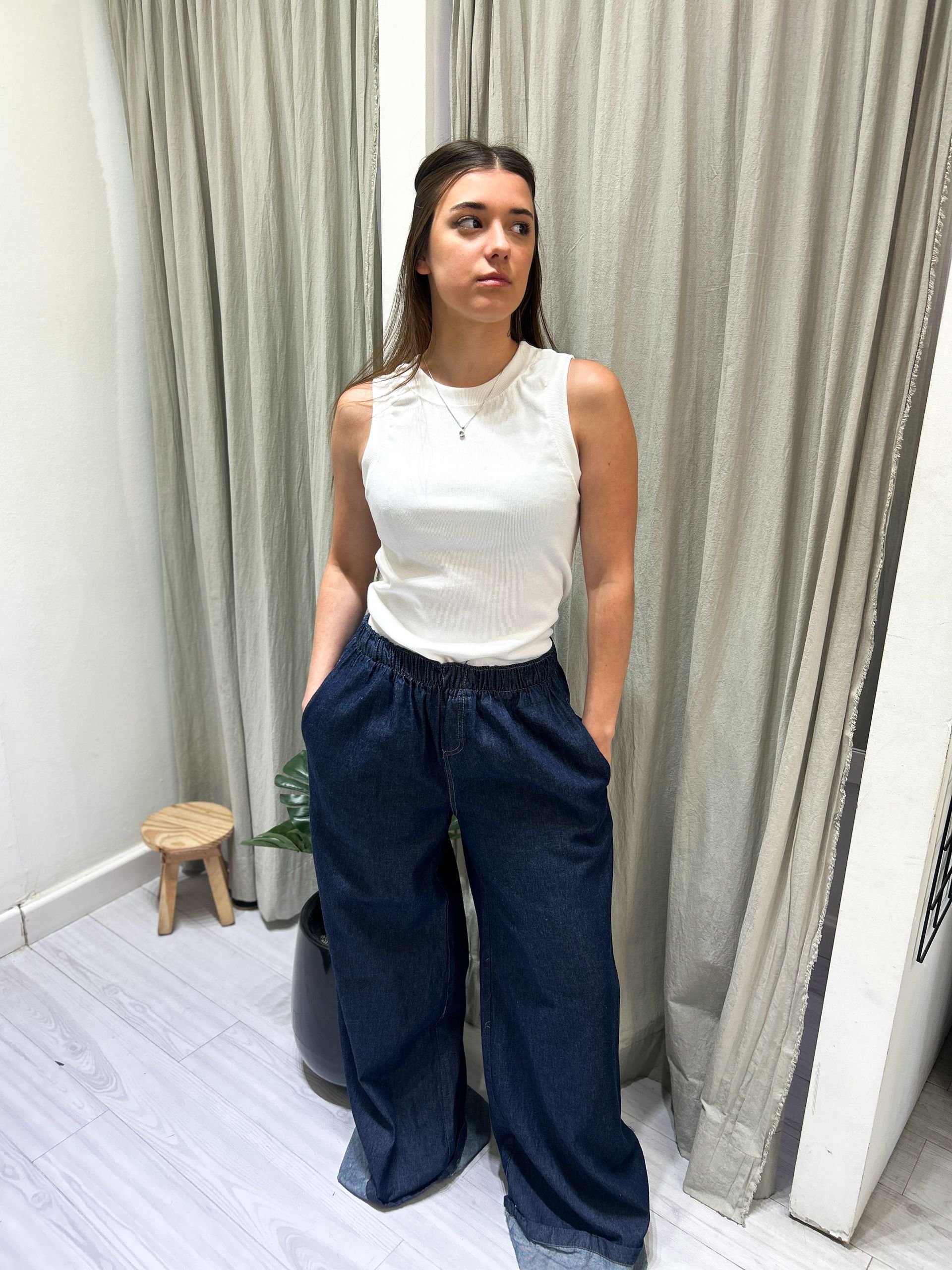 Mujer con camiseta blanca de tirantes y pantalones vaqueros oscuros de pernera ancha, con las manos en los bolsillos.