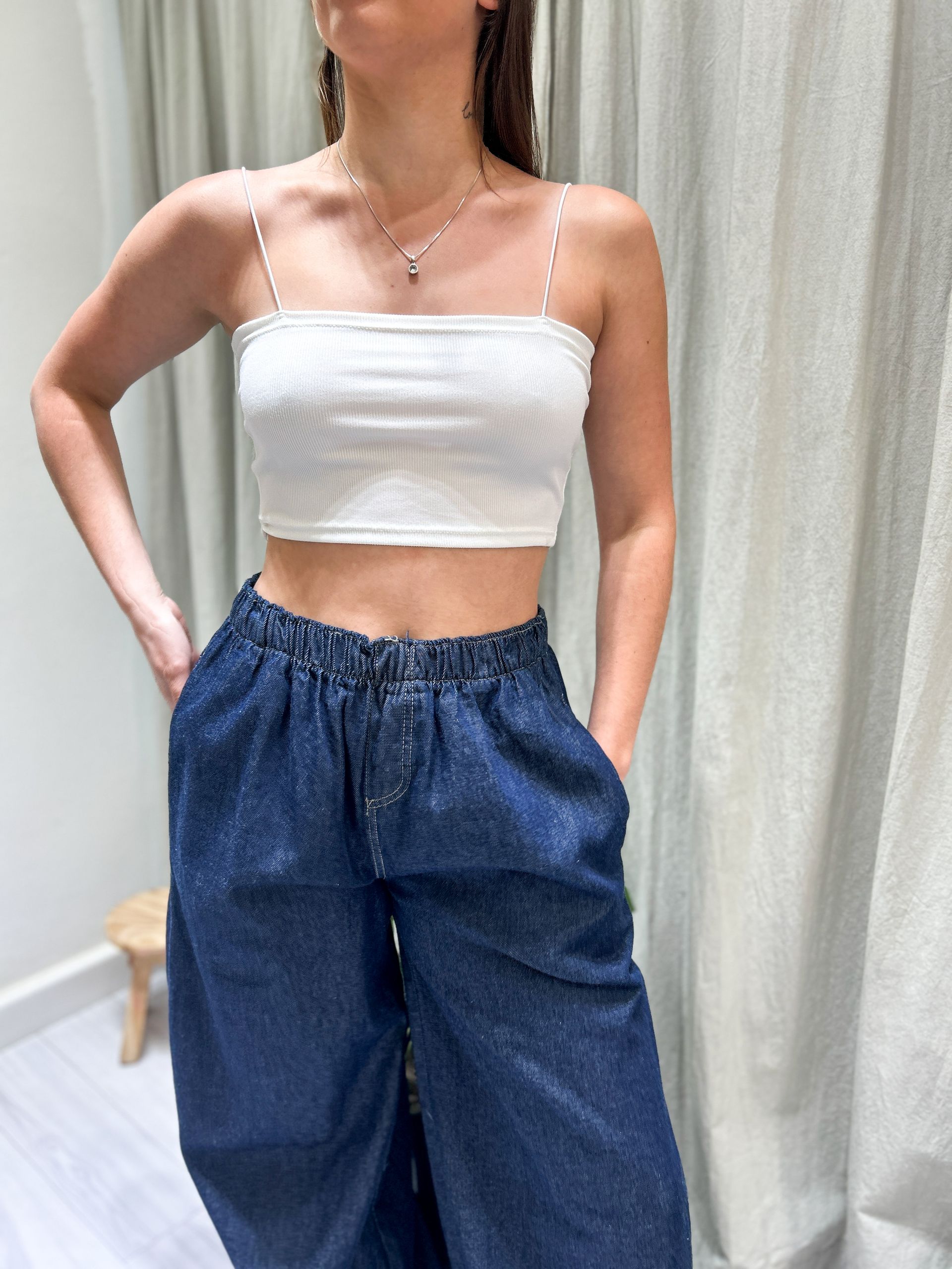 Mujer con camiseta blanca de tirantes y pantalones vaqueros oscuros de pernera ancha, con las manos en los bolsillos.