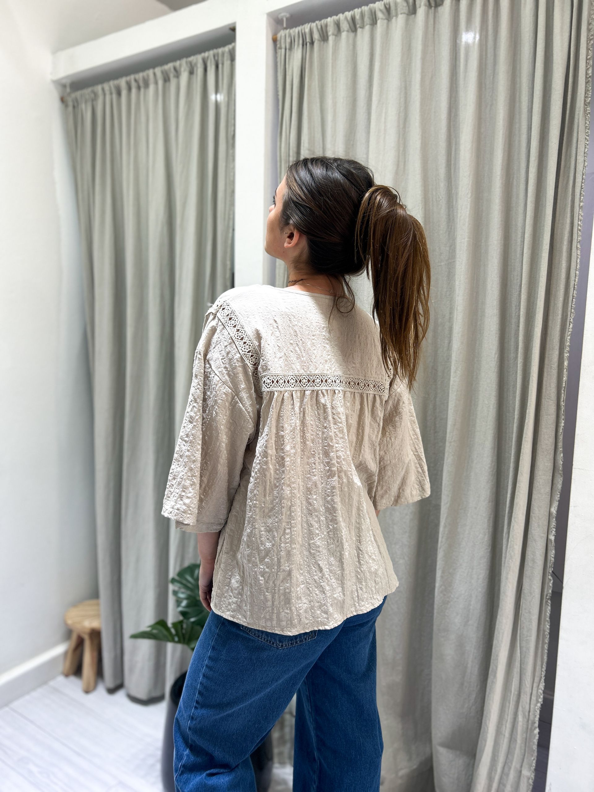 Mujer con blusa negra y jeans sonríe, de pie en el interior con una planta y cortinas.