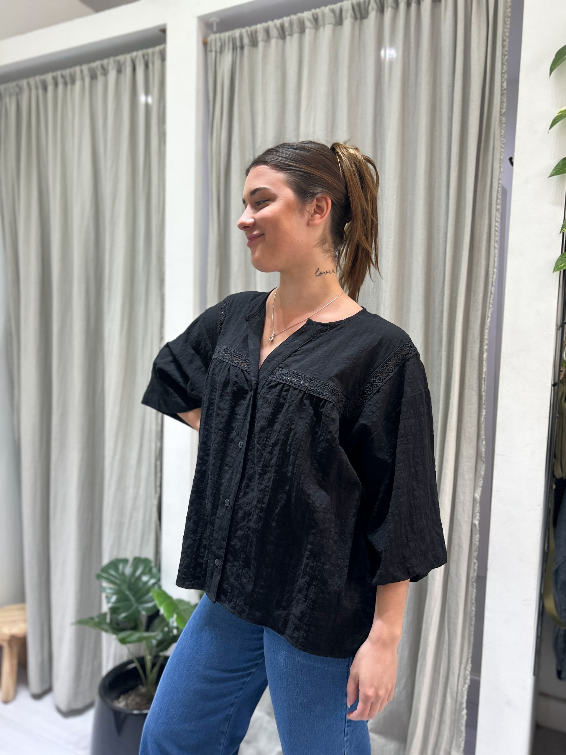 Mujer con blusa negra y jeans sonríe, de pie en el interior con una planta y cortinas.