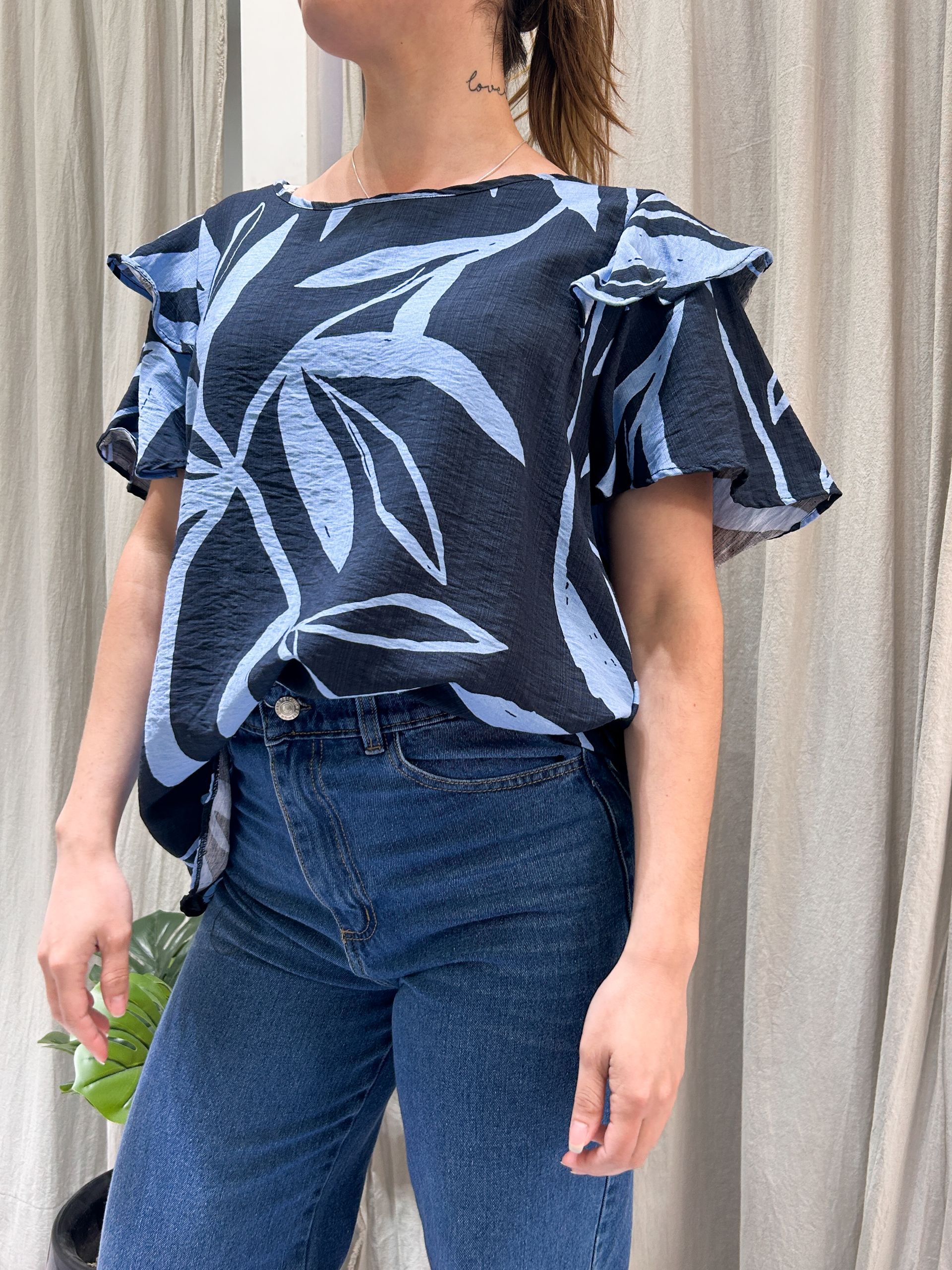 Mujer con blusa azul estampada y jeans, de pie cerca de unas cortinas.