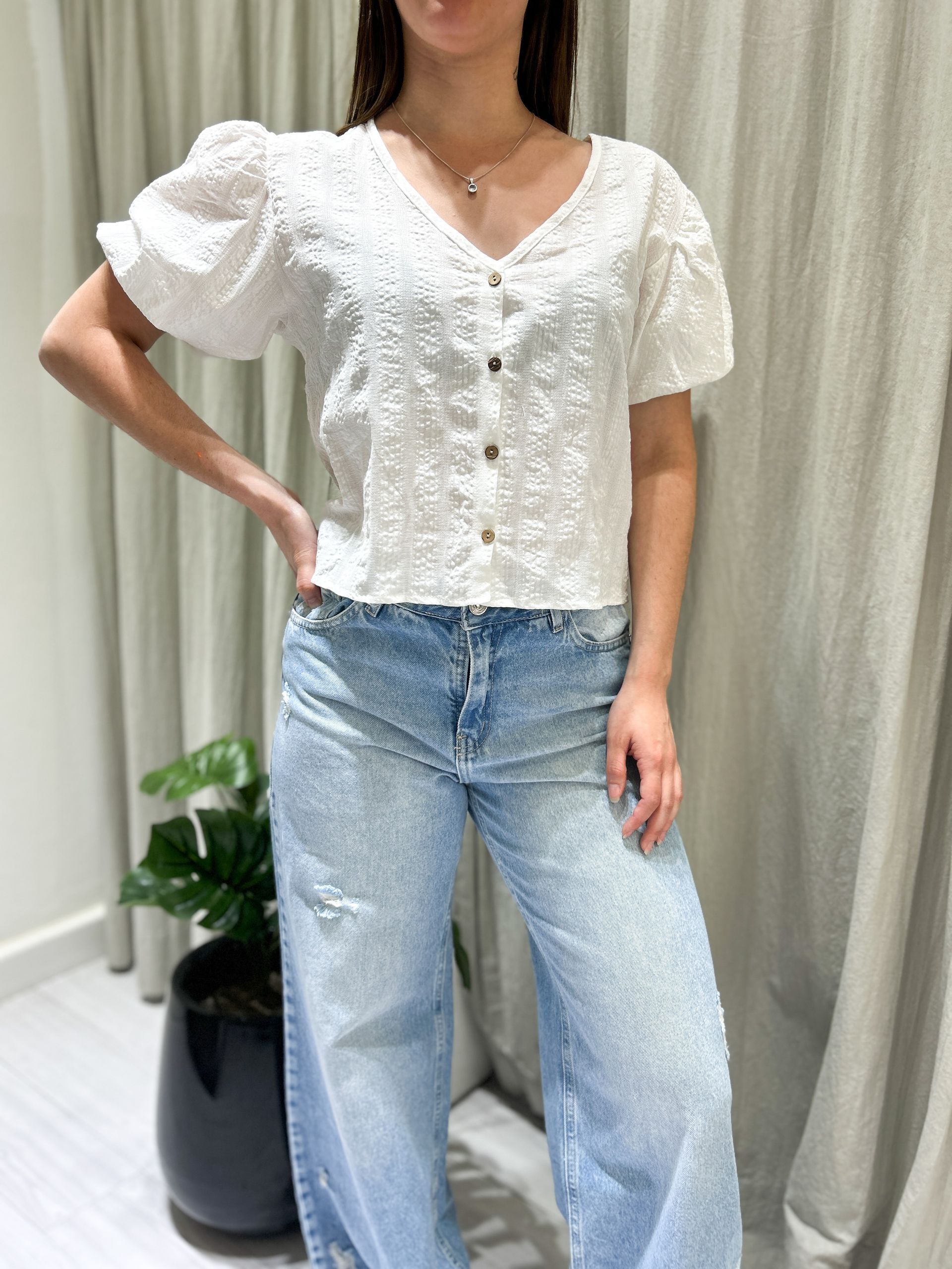 Mujer con blusa blanca bordada y jeans azules, posando cerca de unas cortinas y una planta.
