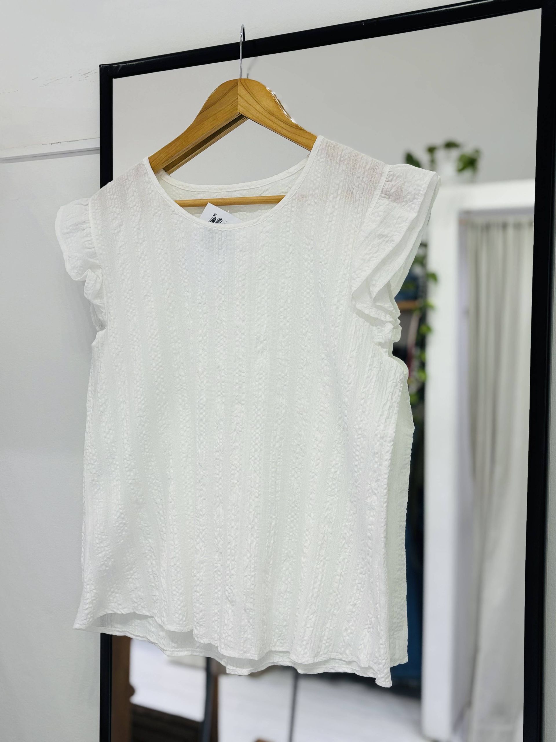 Top blanco texturizado con mangas con volantes colgado en una percha de madera frente a un espejo.