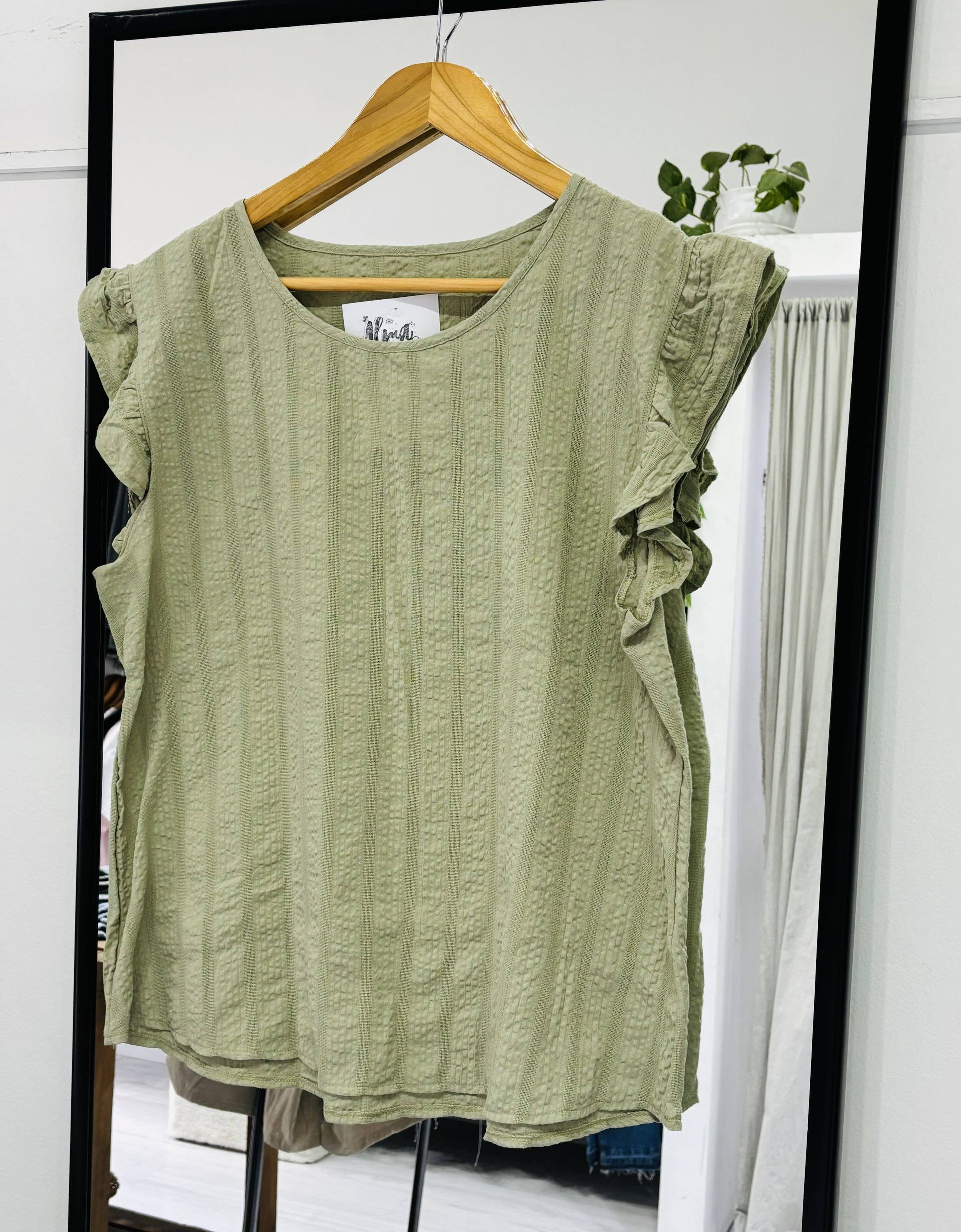 Top verde texturizado con mangas con volantes colgado en una percha de madera frente a un espejo.