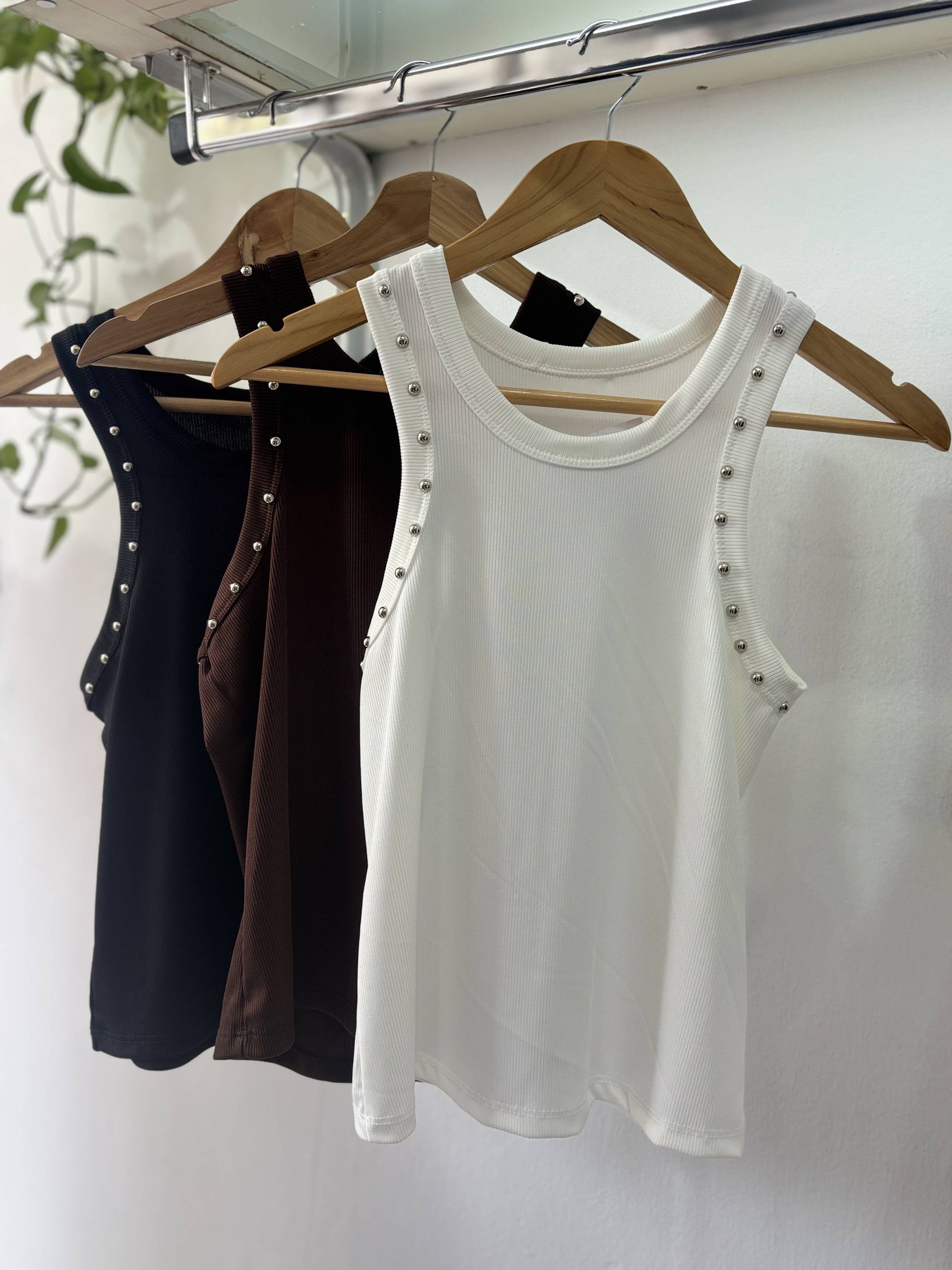 Tres camisetas de tirantes de canalé en negro, marrón y blanco, colgadas en perchas de madera.