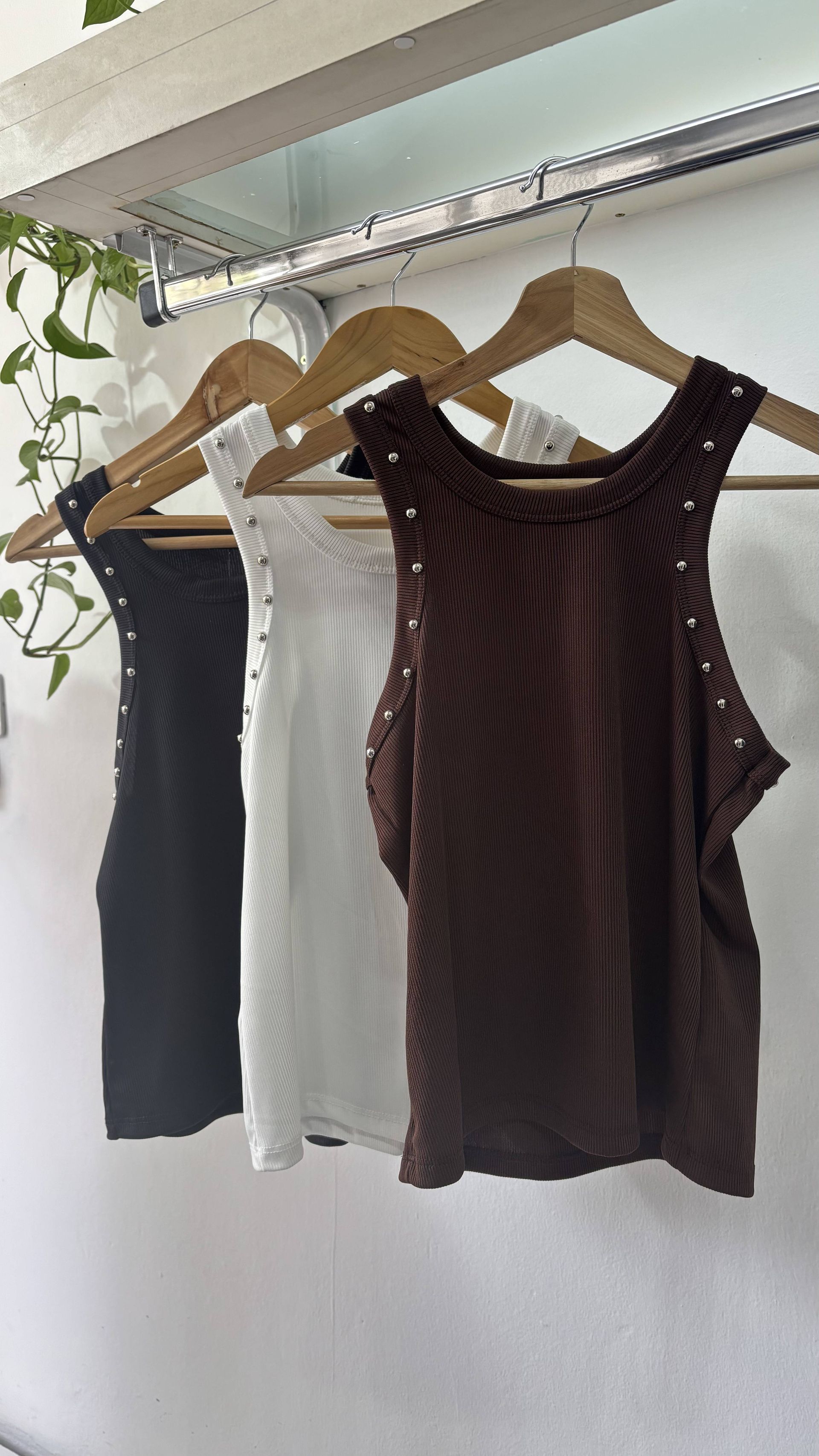 Tres camisetas de tirantes en negro, blanco y marrón colgadas en perchas de madera, contra una pared blanca.