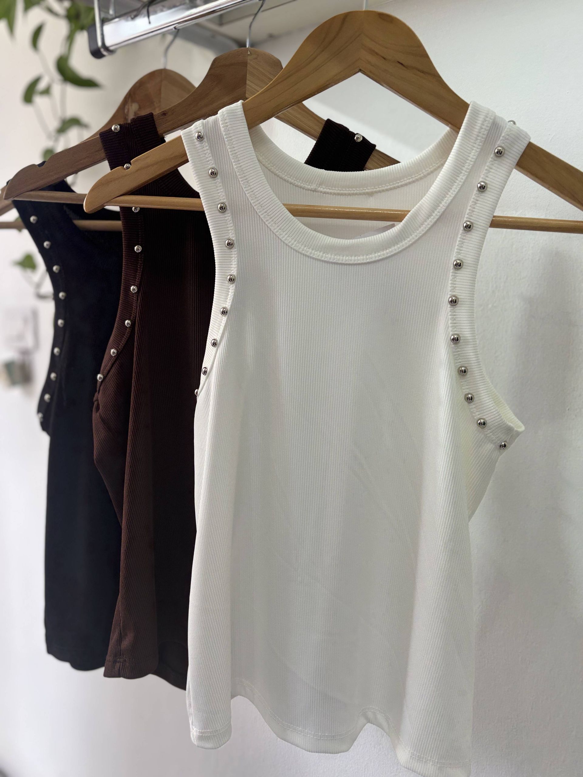 Tres camisetas de tirantes en negro, blanco y marrón colgadas en perchas de madera, contra una pared blanca.