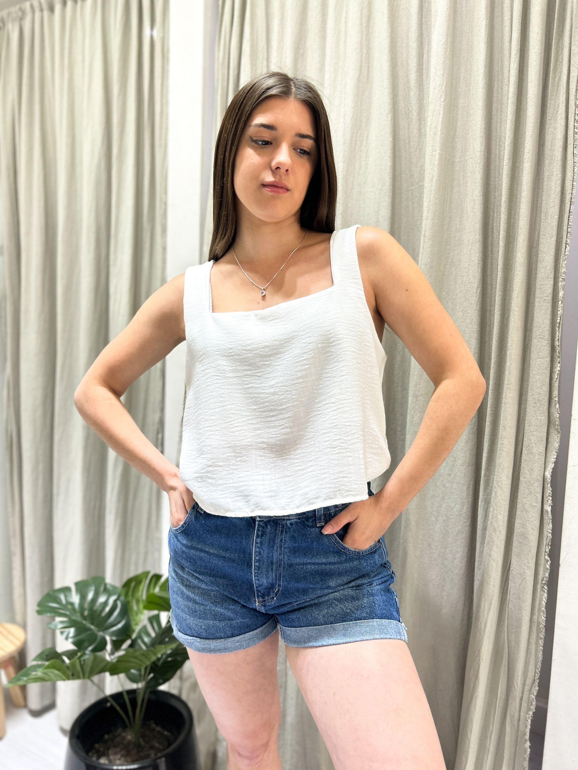 Mujer con blusa blanca y pantalones cortos vaqueros, manos en los bolsillos, posando en interiores.