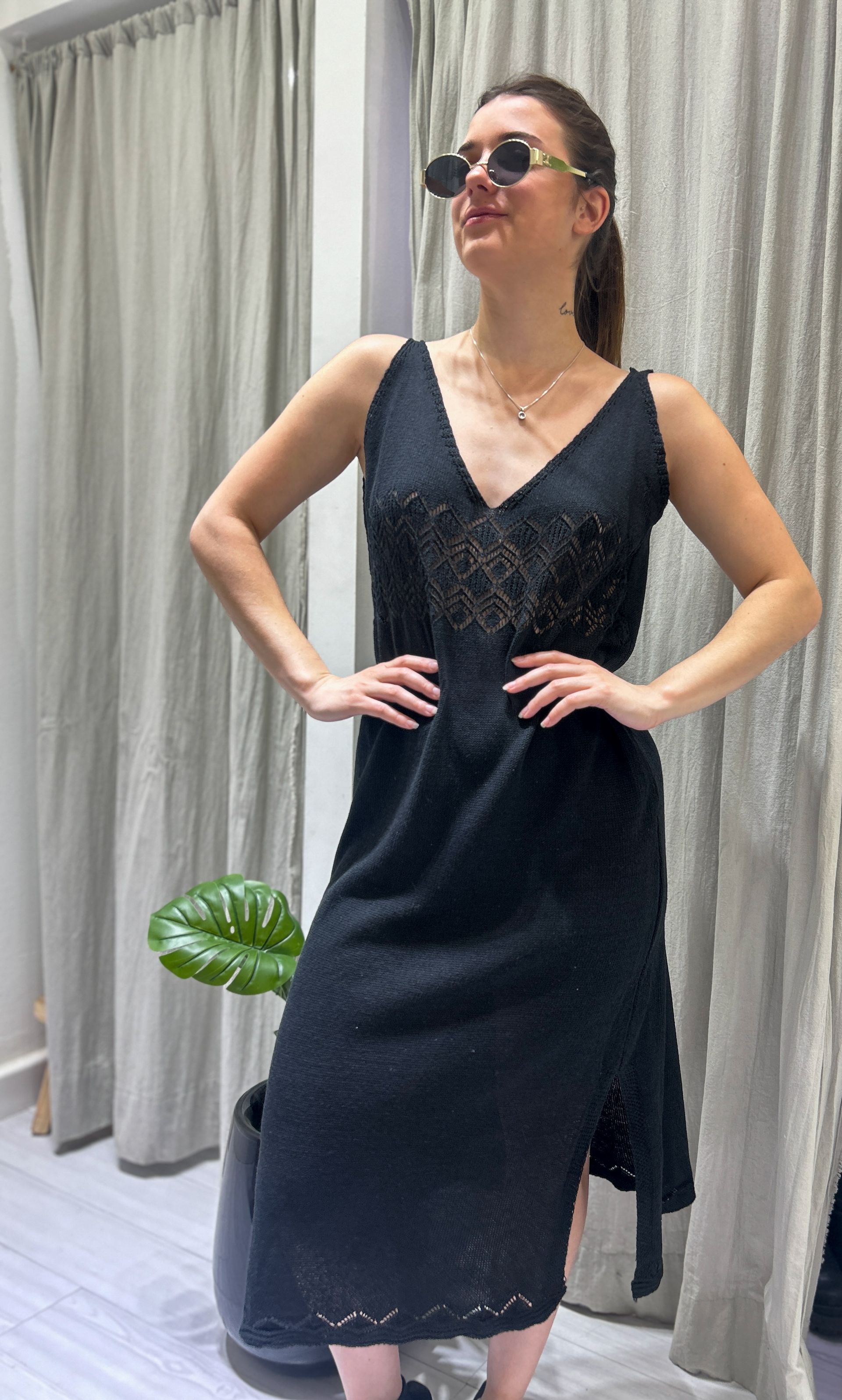 Mujer con vestido negro y gafas de sol, posando en interiores con las manos en las caderas.