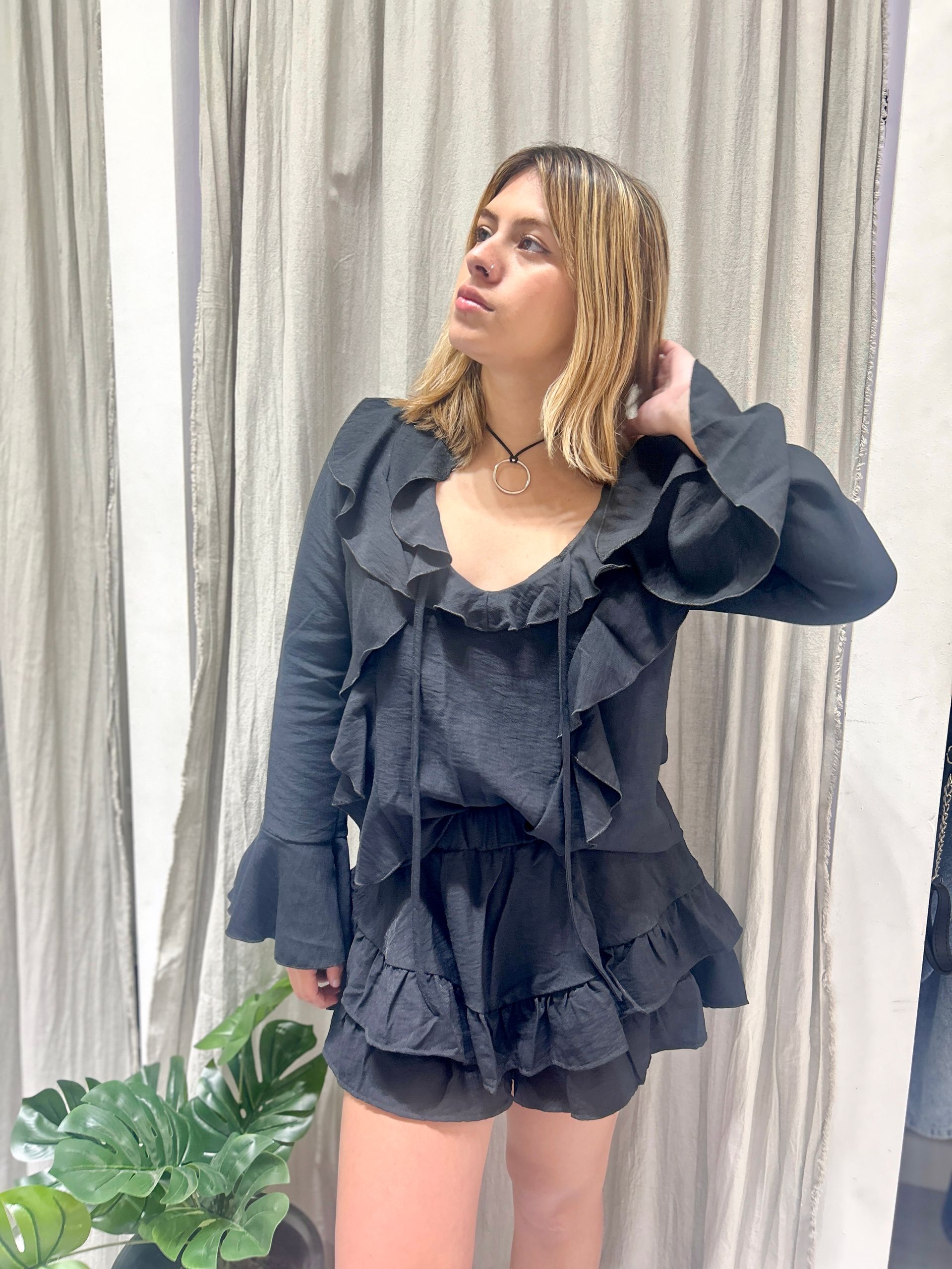 Mujer con atuendo oscuro con volantes mirando hacia arriba, en interiores cerca de una cortina y vegetación.