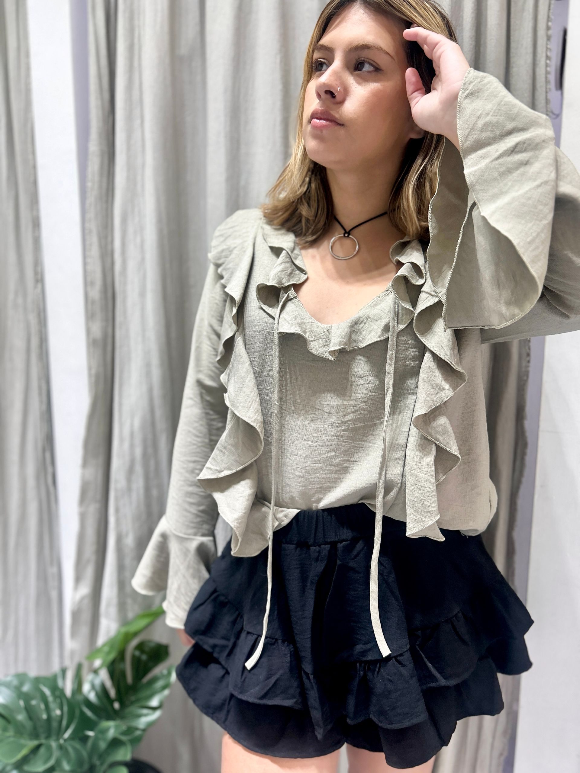 Mujer con atuendo oscuro con volantes mirando hacia arriba, en interiores cerca de una cortina y vegetación.