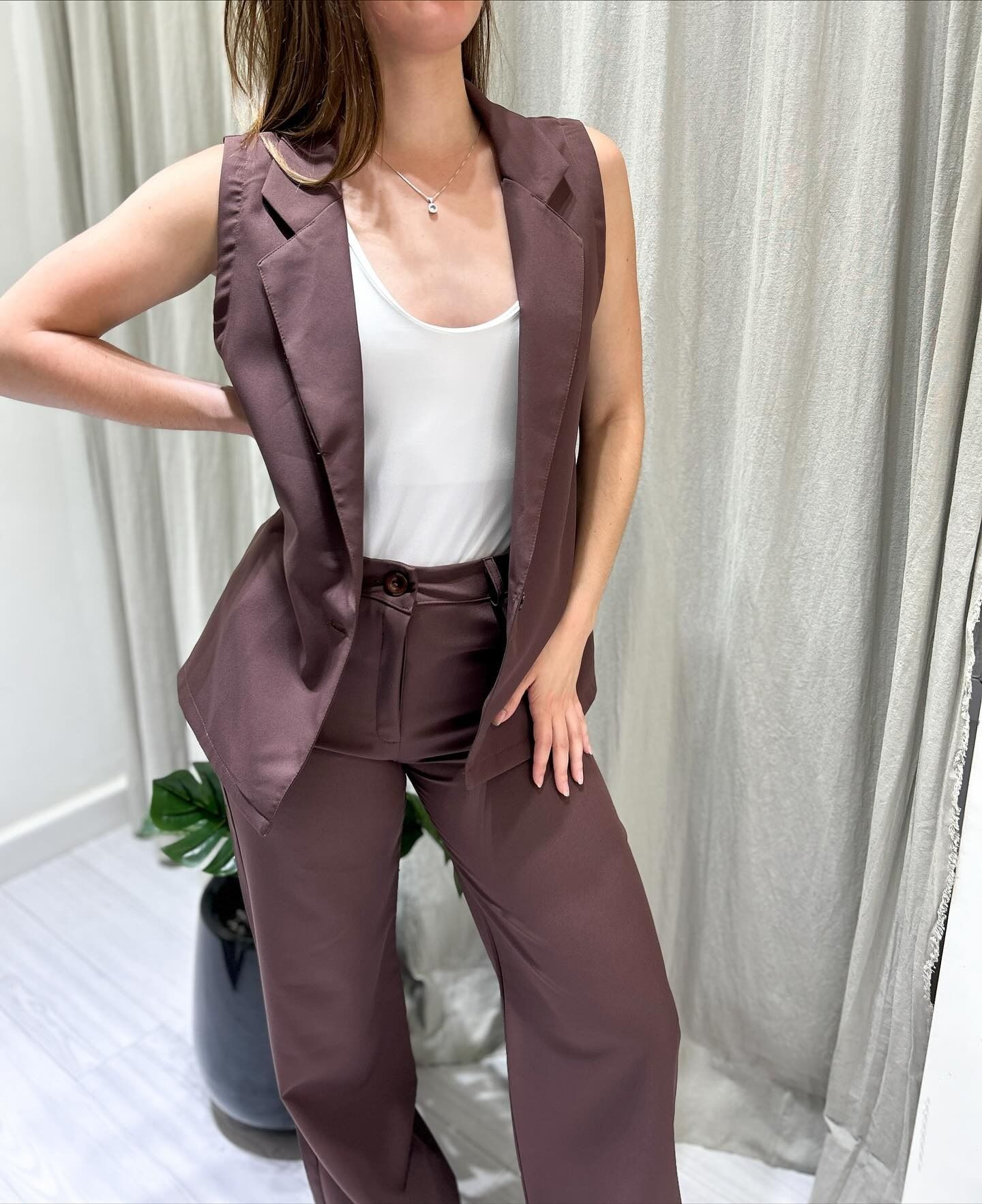 Mujer con chaleco y traje pantalón marrón, sobre una camiseta blanca de tirantes, de pie en el interior.