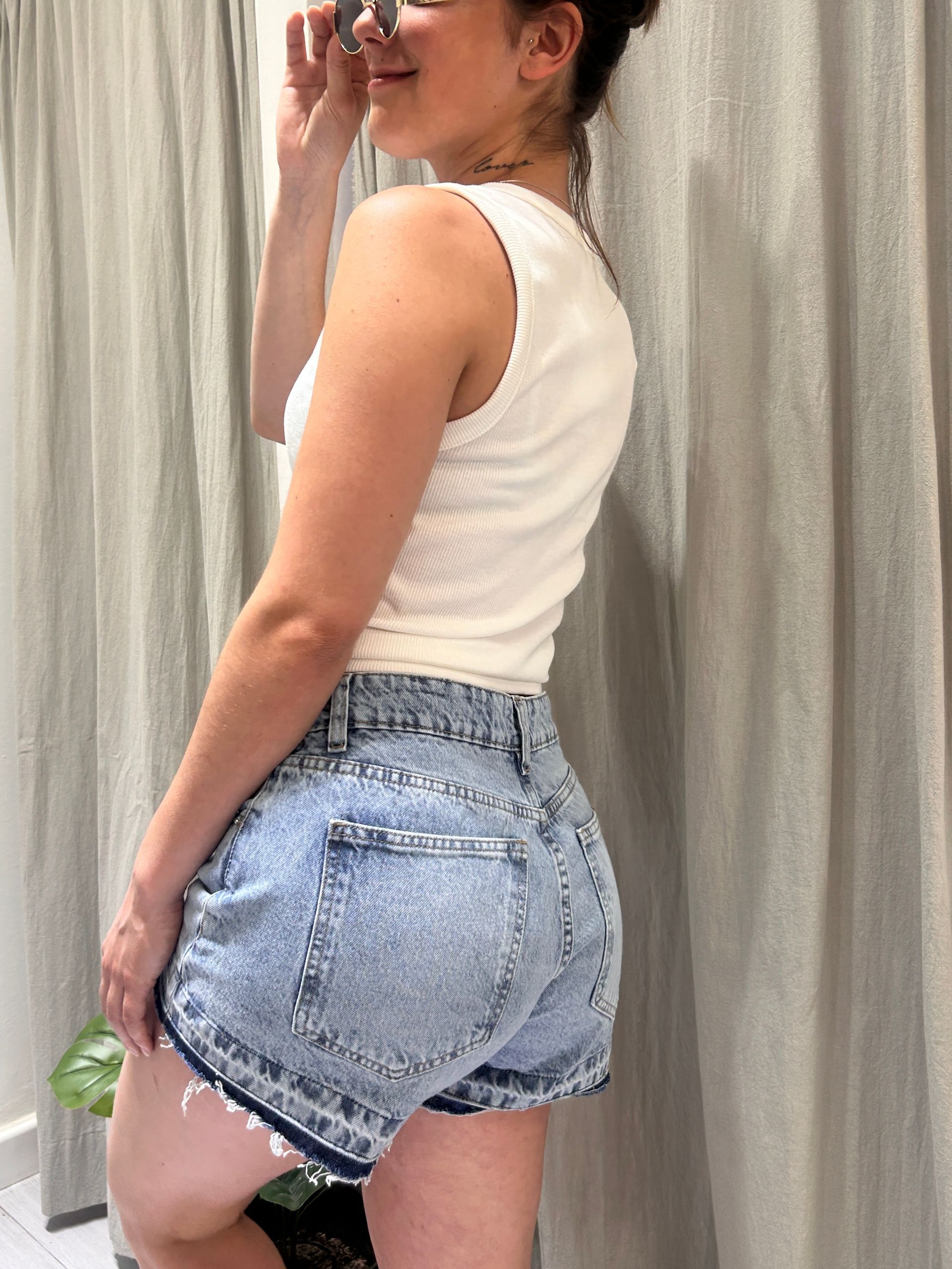 Mujer con camiseta blanca de tirantes y pantalones cortos vaqueros, manos en los bolsillos, de pie en el interior.