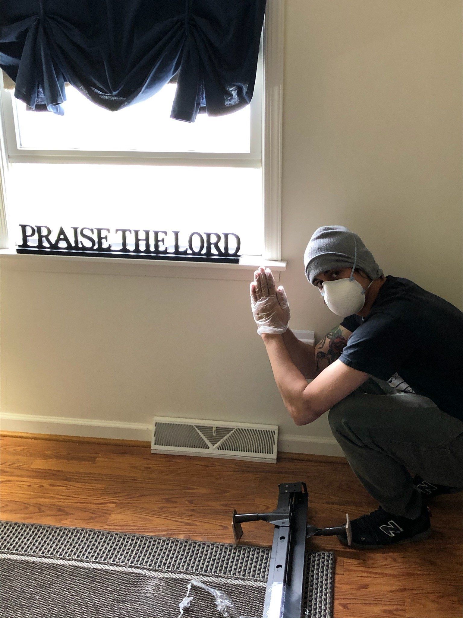A man wearing a mask is squatting down in front of a window with a sign that says praise the lord.