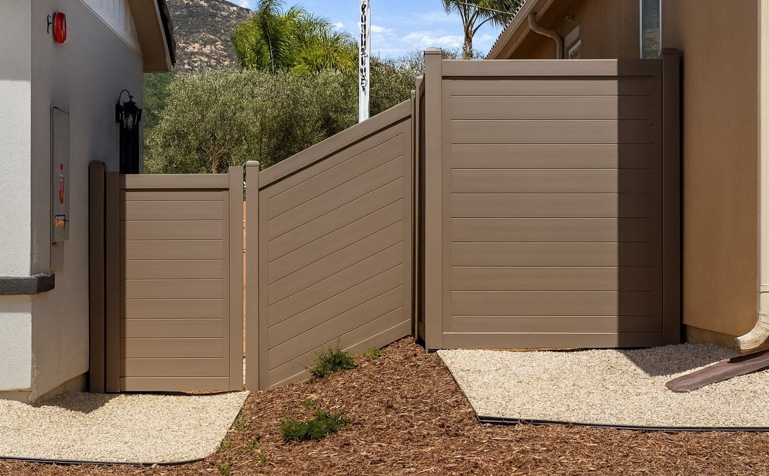 A brown horizontal slat fence and gate connecting two beige exterior walls over a landscaped ground of mulch and gravel.